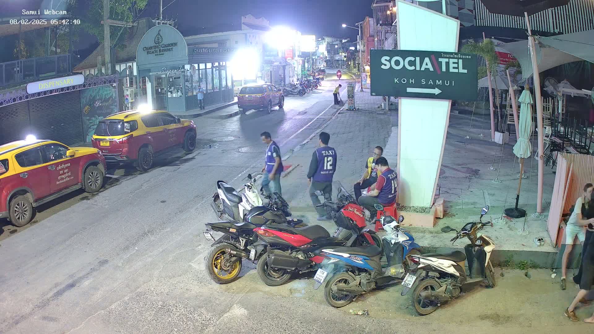 At night, several people stand near parked motorcycles and taxis on a street in front of buildings and outdoor seating areas.