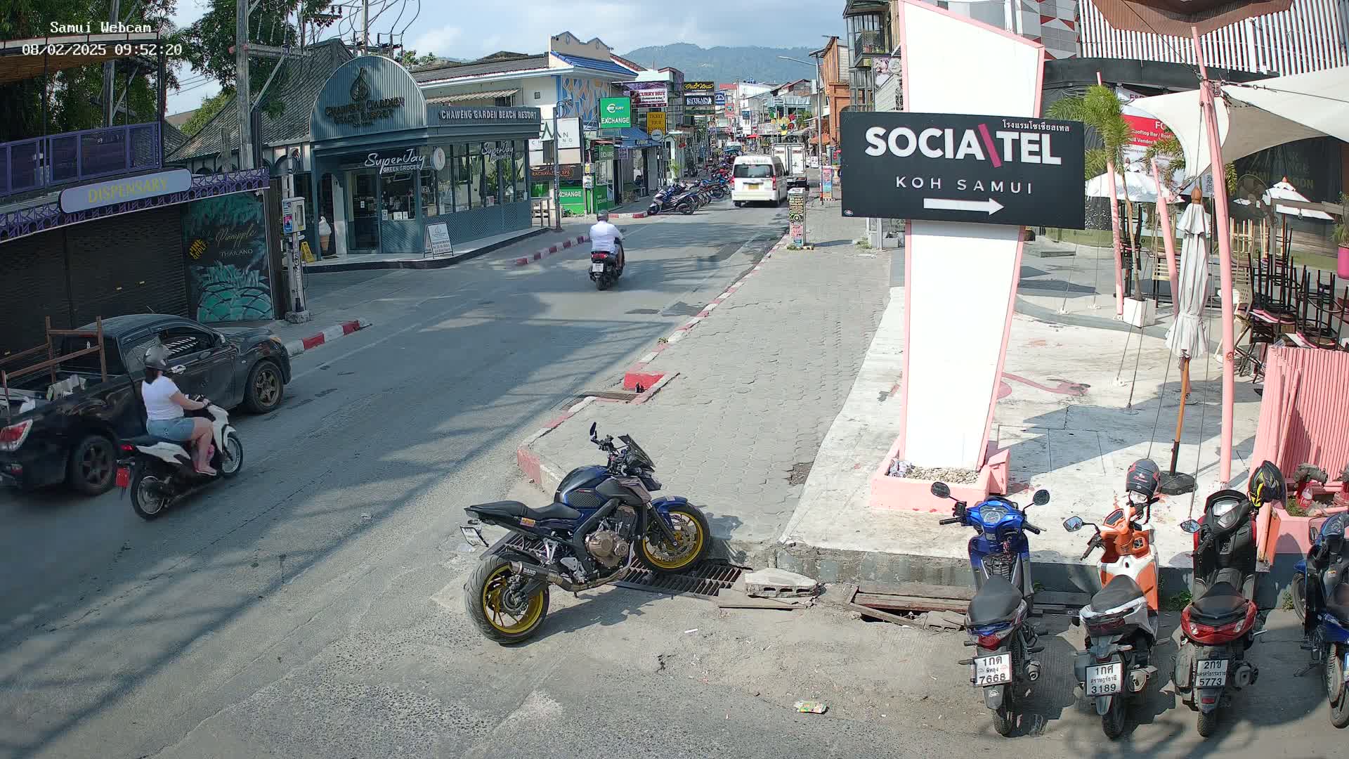 A sunny street scene shows several motorbikes and scooters parked and in motion, along with a pickup truck and a few pedestrians, in front of shops and businesses.