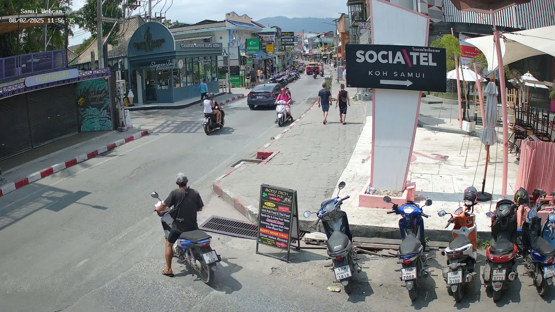 A sunny street scene shows several people walking and riding motorbikes, with parked motorbikes lining the sidewalk near outdoor seating under pink umbrellas.