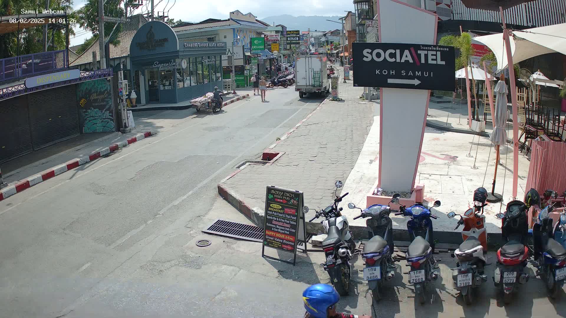 A sunny street scene shows a row of parked motorbikes, pedestrians, and shops, with a truck parked further down the road.