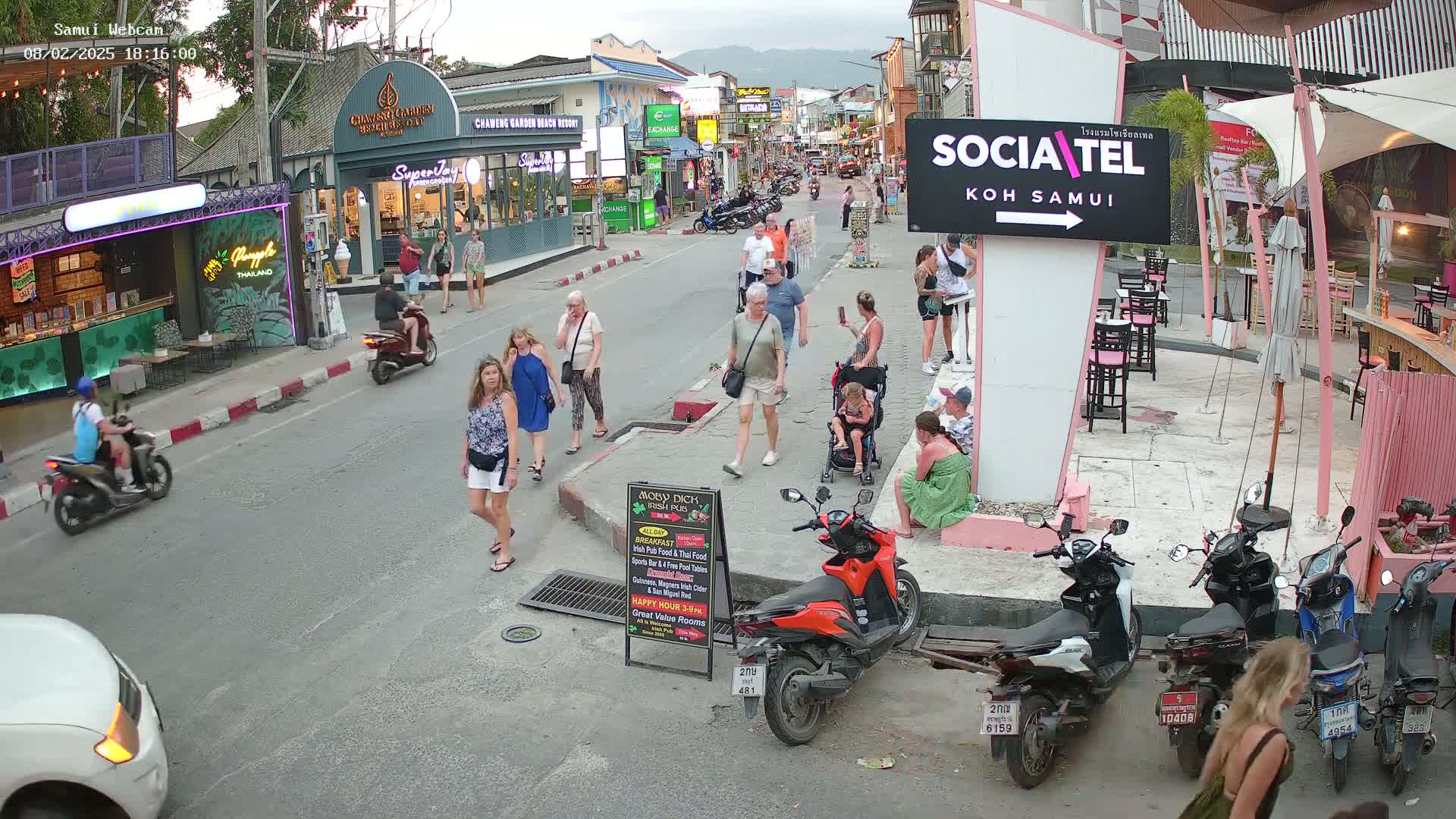 A street scene at dusk shows pedestrians and motorbikes parked along a road lined with shops and restaurants.