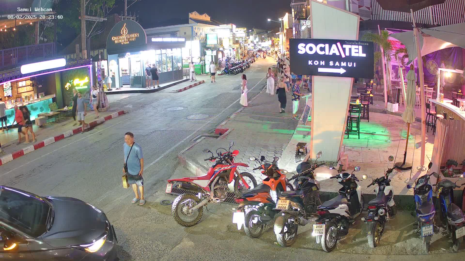 At night, a street scene shows several people walking along a road lined with shops and restaurants, with parked motorcycles and a car visible.