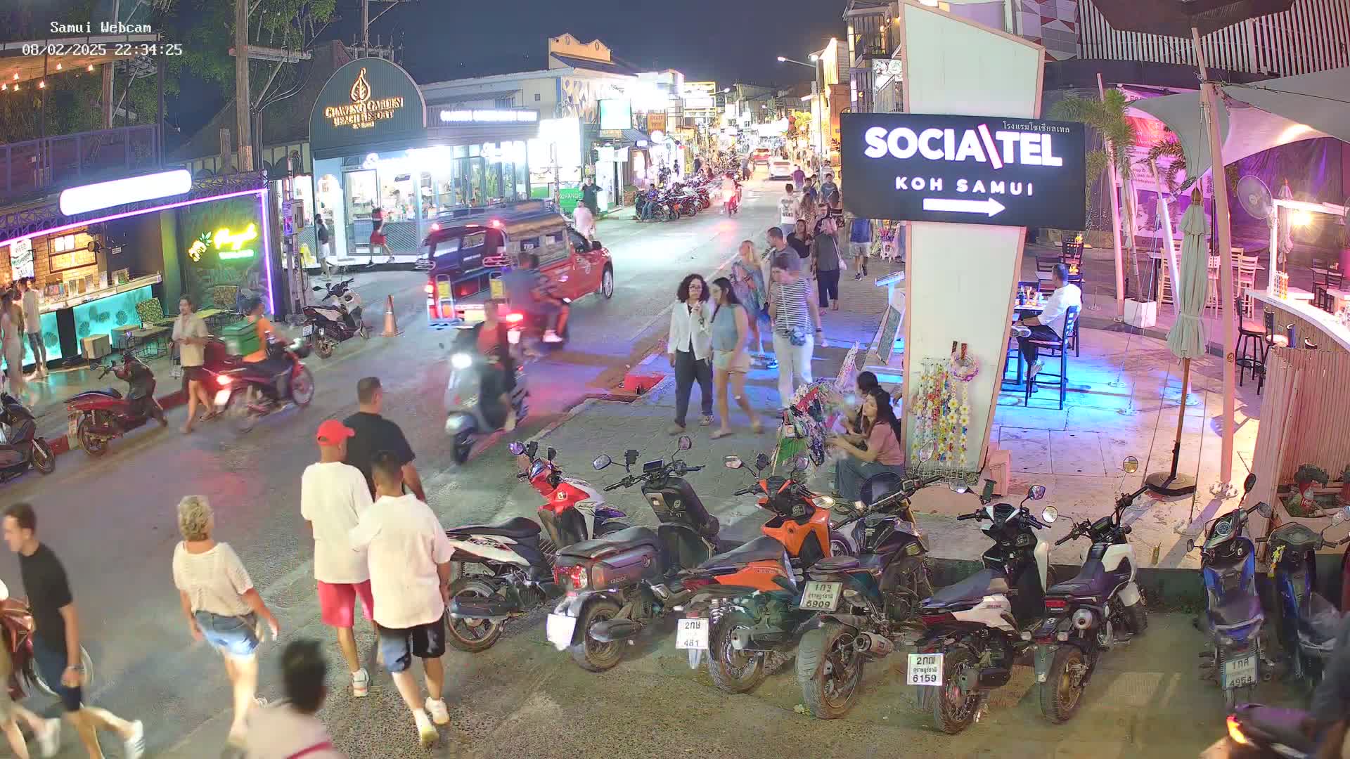 A nighttime street scene shows many people walking among parked motorcycles and several shops, under a clear sky.