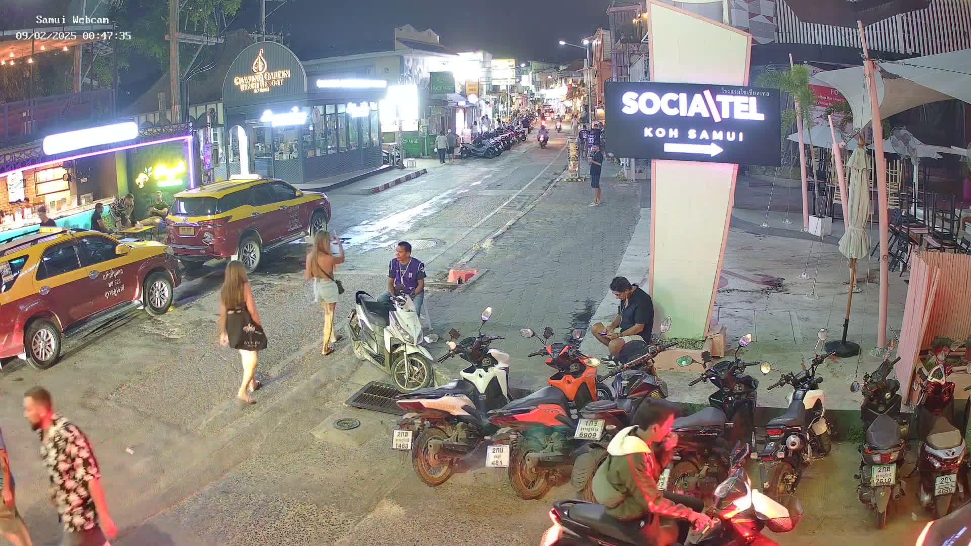 A nighttime street scene shows several people walking and standing near parked taxis and motorbikes, under partly cloudy skies.