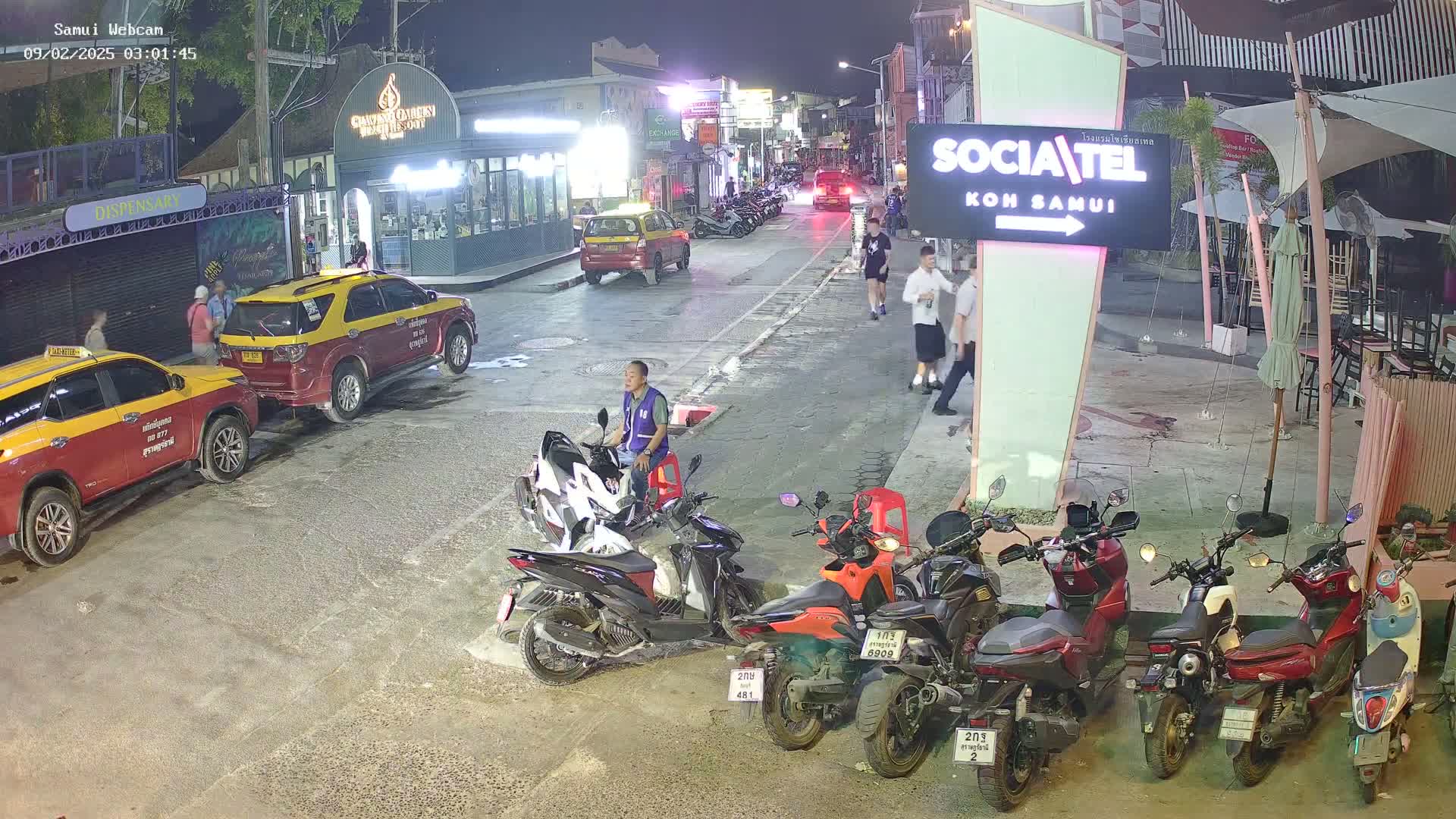 At night, several parked motorcycles and taxis line a street with pedestrians walking past outdoor seating areas under a partly cloudy sky.