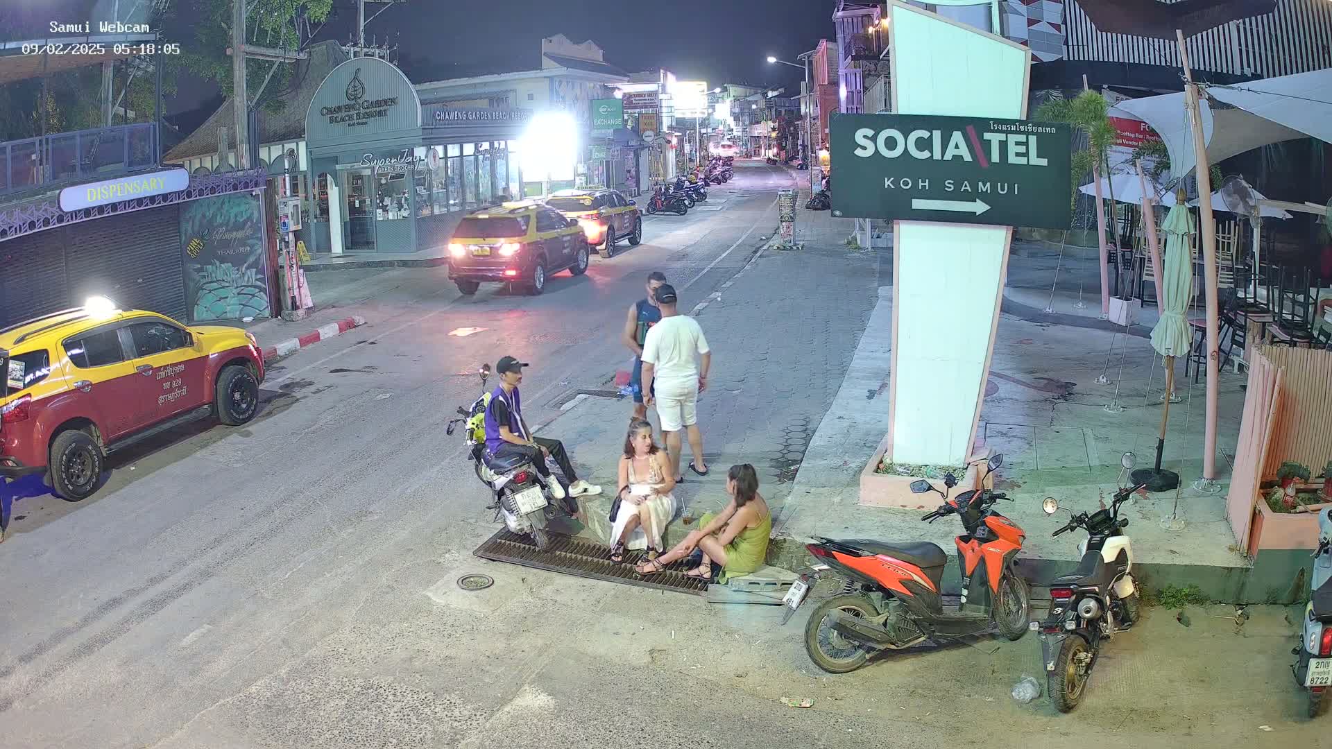 At night, several people are sitting and standing on a street near parked cars and motorcycles, under outdoor lighting.