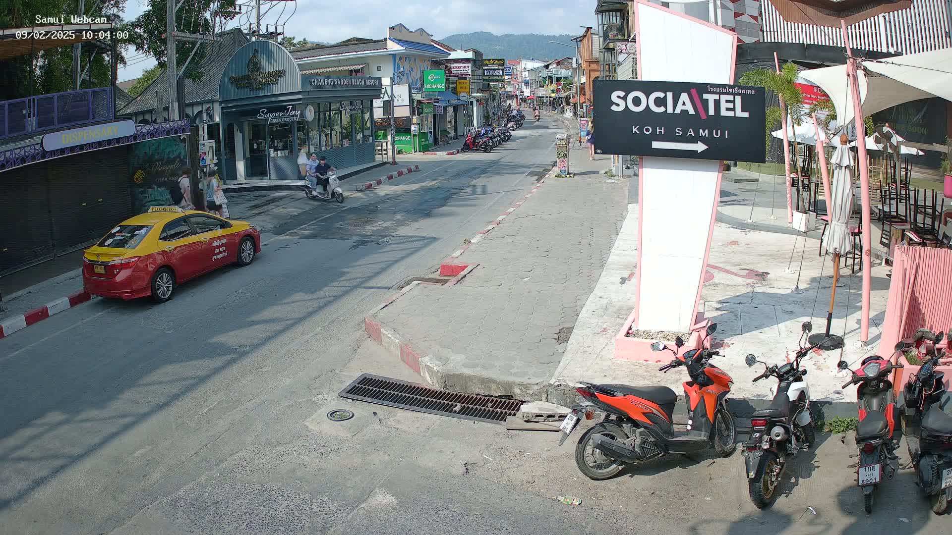 A sunny street scene shows parked motorbikes, a yellow taxi, and pedestrians near shops and restaurants.
