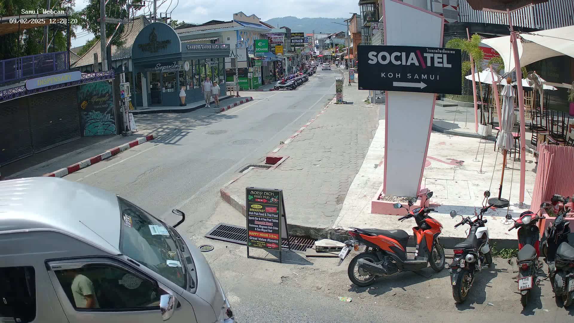 A sunny street scene shows several parked motorbikes, a silver van, and pedestrians walking past shops and restaurants.