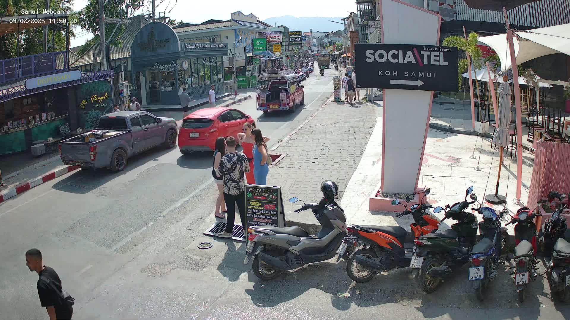 A sunny street scene shows several people standing together near parked cars and motorbikes, with shops and restaurants lining the street.