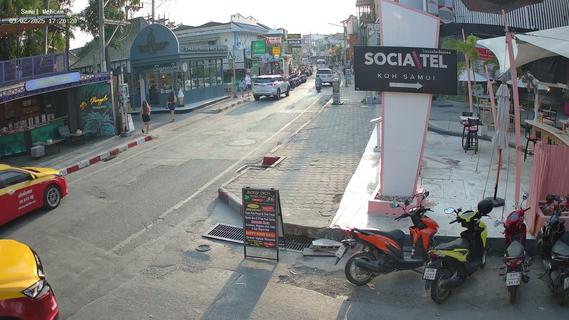 A street scene in bright sunlight shows several pedestrians, cars, and parked motorbikes, with shops and restaurants lining the street.