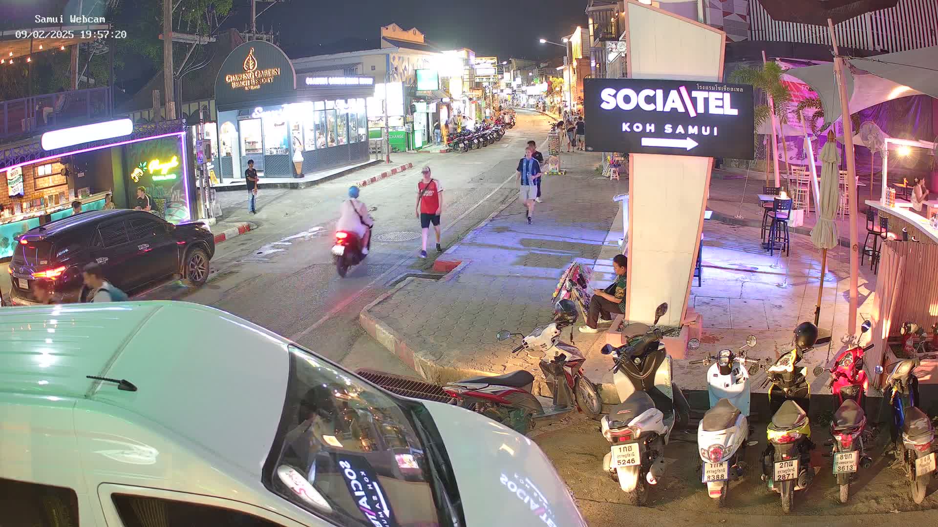 A night scene of a street with parked motorbikes, pedestrians, and vehicles, under nighttime lighting conditions.