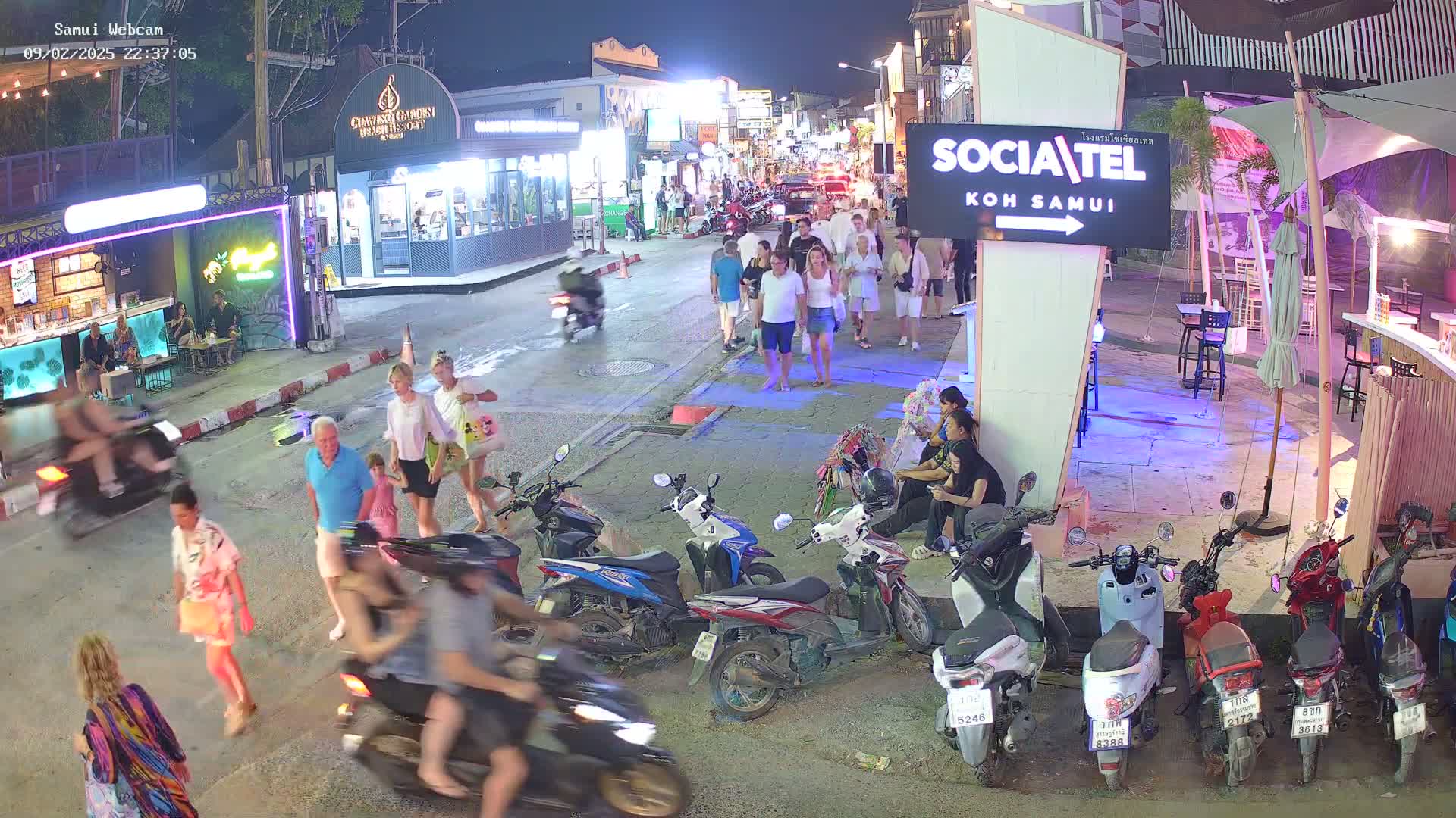 A nighttime street scene shows pedestrians and motorcyclists moving about, with parked motorcycles lining the side of a road next to several businesses and outdoor seating areas.