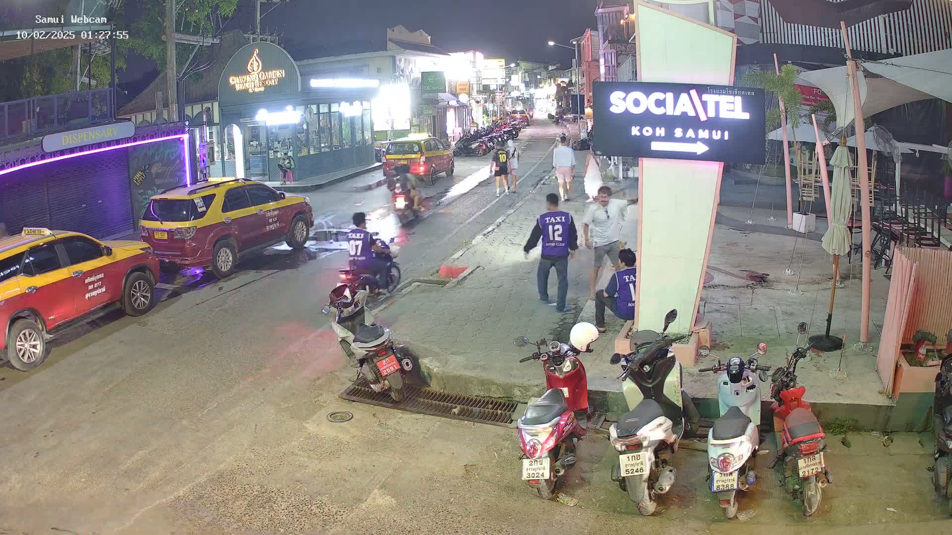 A nighttime street scene shows several people standing around near parked taxis and motorbikes, with some people on motorbikes moving along a road lined with shops and restaurants under well-lit conditions.