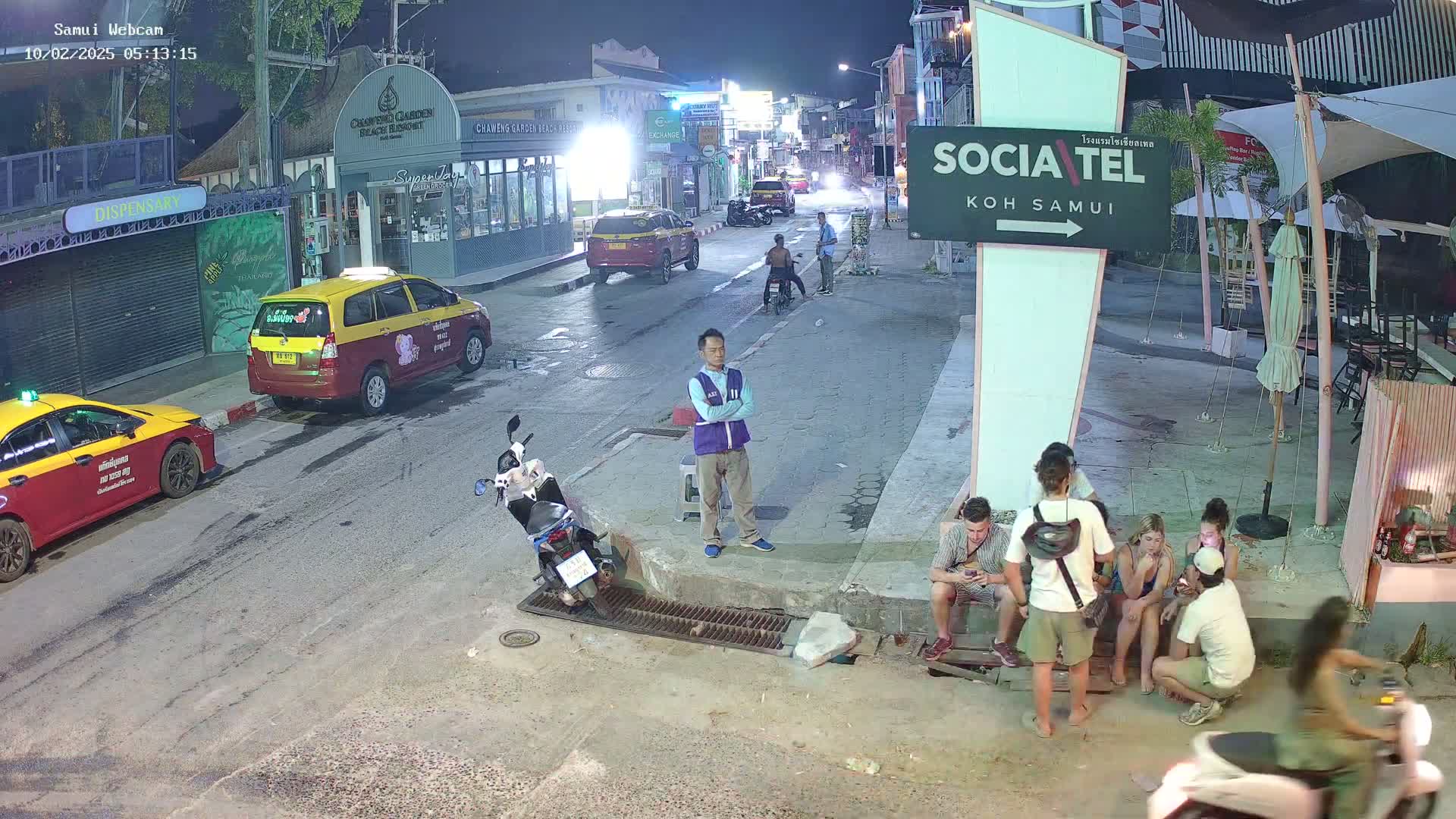 At night, on a street with parked taxis and motorbikes, several people are gathered near a restaurant, while others walk along the road.