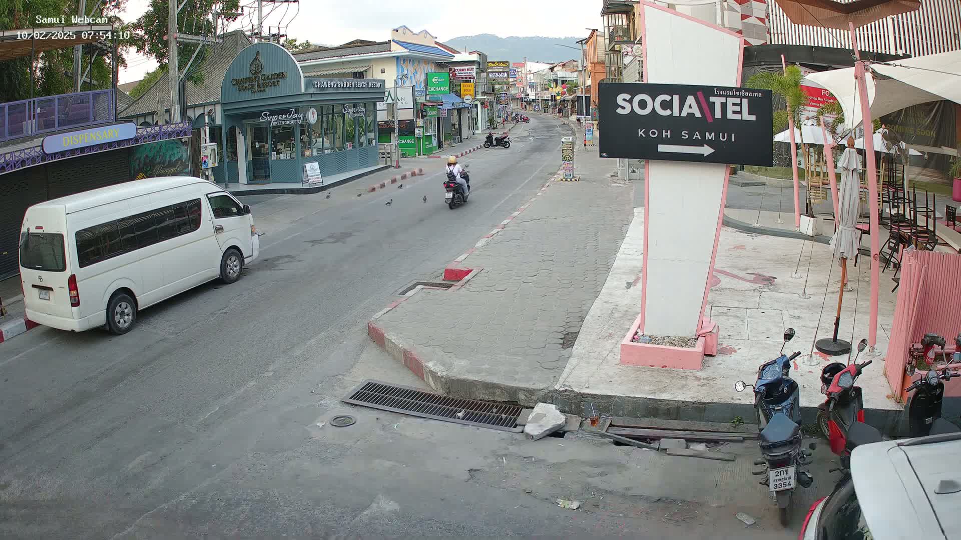 A mostly empty street scene in Koh Samui, Thailand, shows a white van, motorbikes, and pedestrians under an overcast sky.