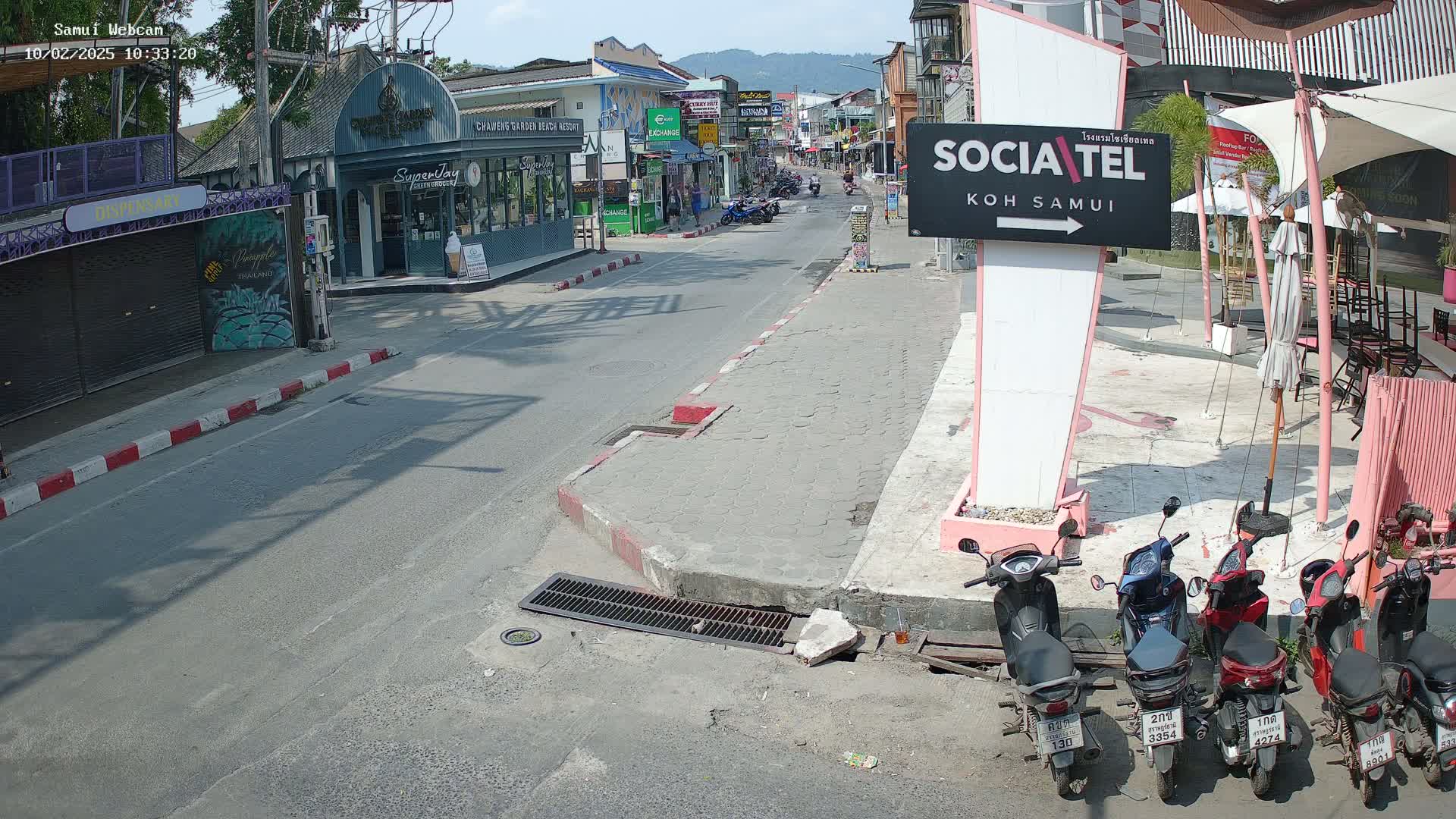 A sunny street scene shows a road with parked motorbikes, businesses lining the street, and a few people walking in the distance.