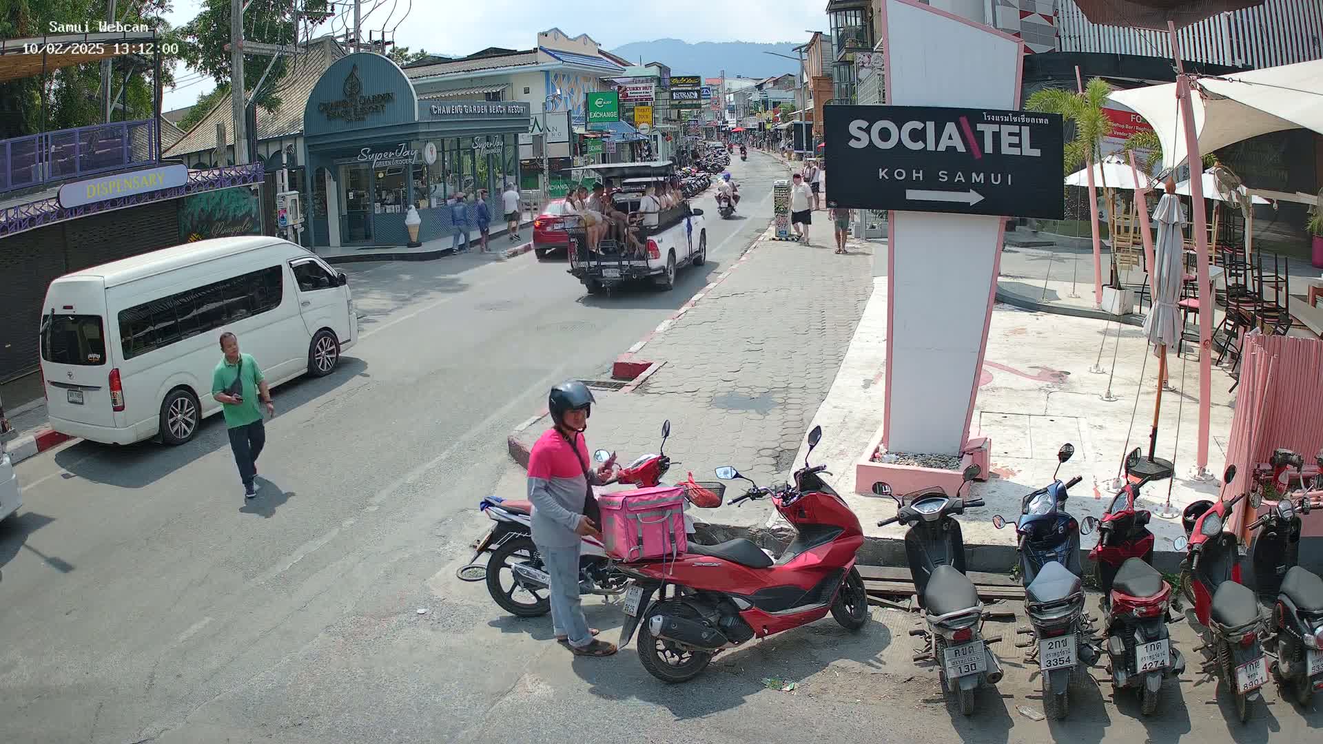 A sunny street scene shows pedestrians, vehicles including a van and a tourist-style open-air vehicle, and several parked motor scooters, with buildings and businesses lining the street.