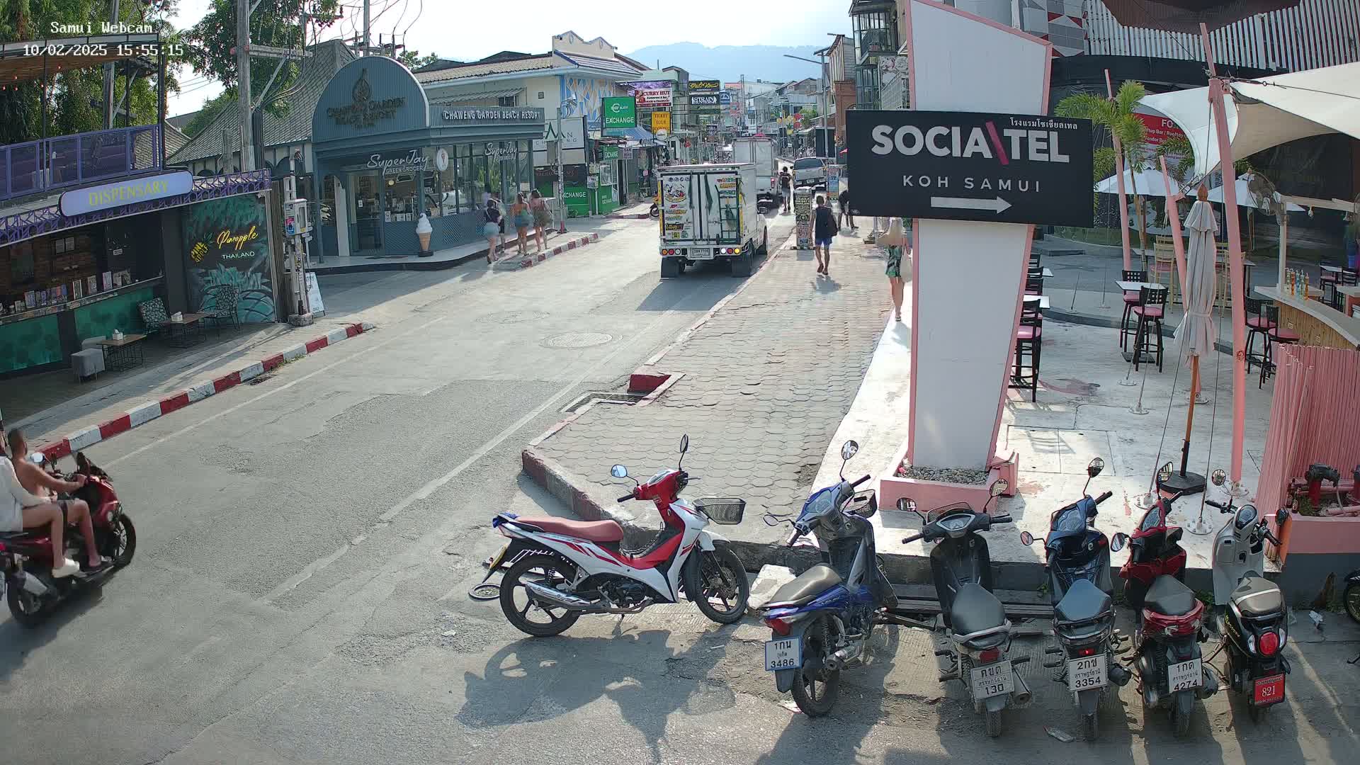 A sunny street scene shows several parked motorbikes, a few pedestrians, and a delivery truck, with buildings and shops lining both sides of the road.