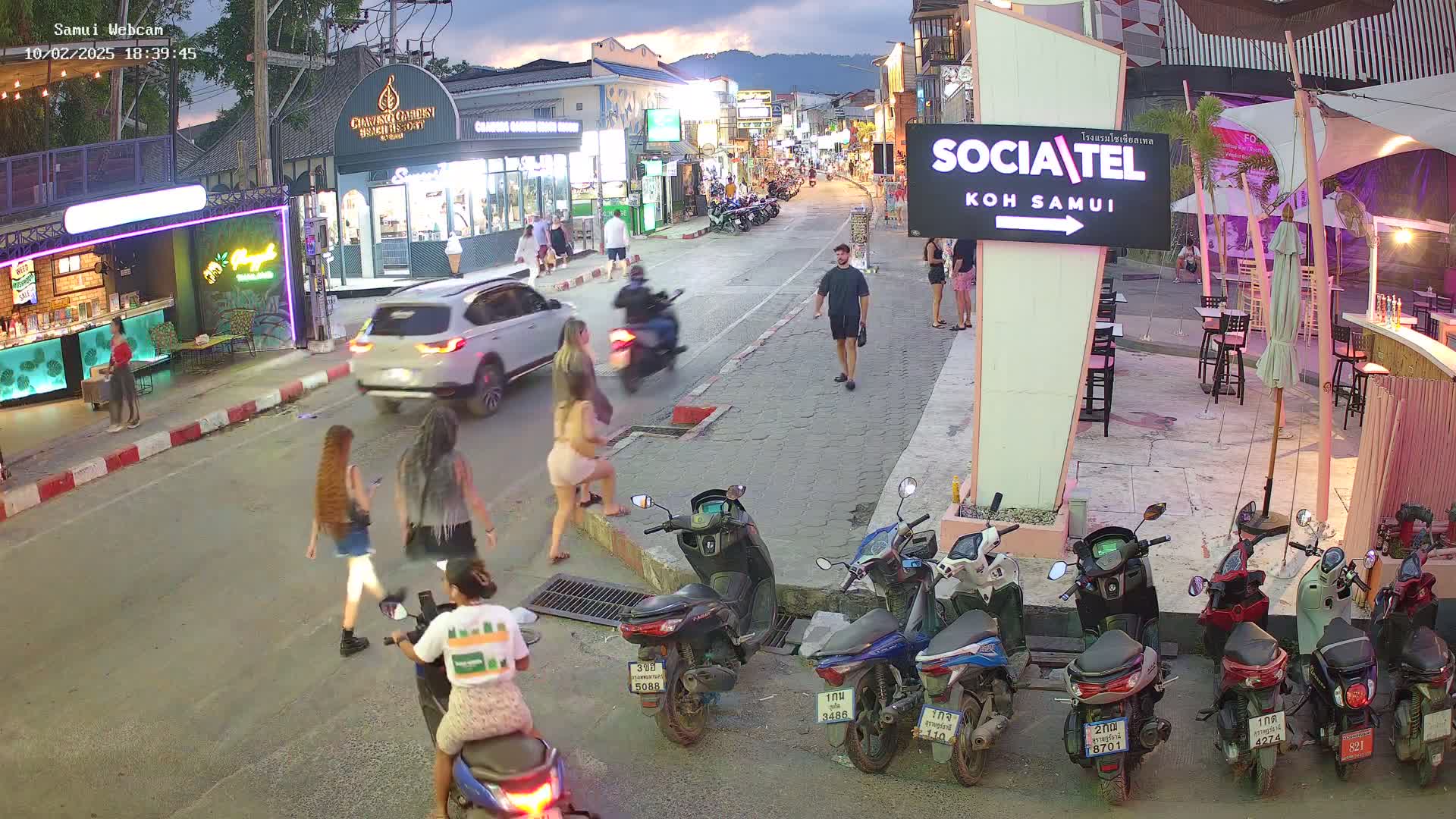 A twilight street scene shows pedestrians and motorbikes in a town, with several shops and restaurants visible along the sides of the road.