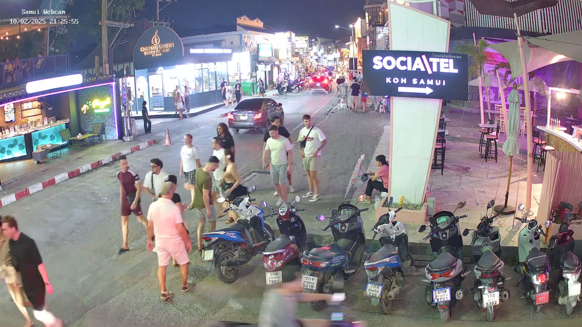 A nighttime street scene shows people walking and standing near shops and restaurants, with several parked motorbikes and cars, under clear night skies.