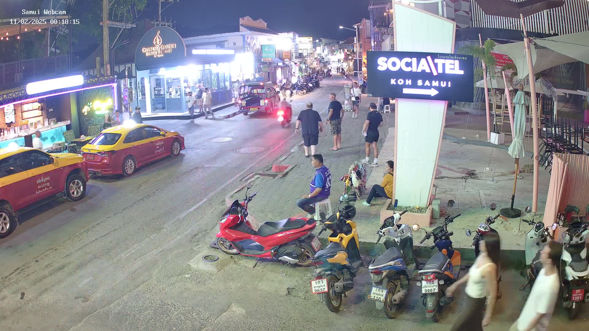 A nighttime street scene shows several people walking and standing near parked motorcycles and taxis, under outdoor lights.