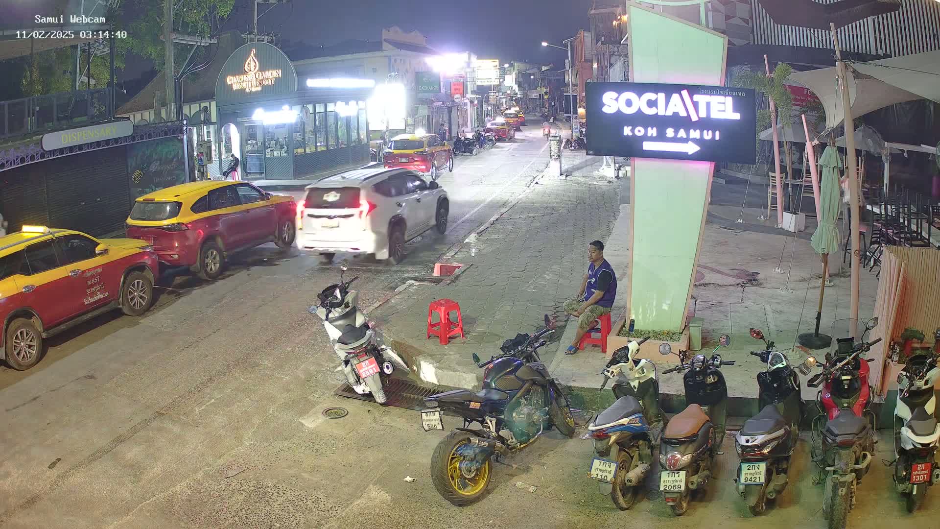 A nighttime street scene shows parked cars and motorcycles, with a man sitting on a stool near a sign, under dark, clear skies.