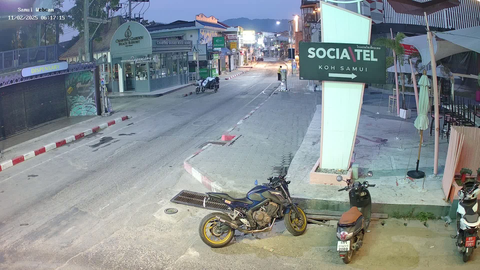 A mostly empty street at twilight, lined with shops and restaurants, with a few parked motorcycles and a scooter.