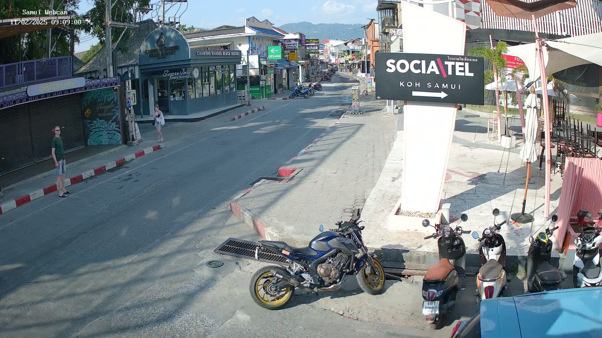 A sunny street scene shows a few pedestrians and parked motorcycles in front of shops and restaurants.