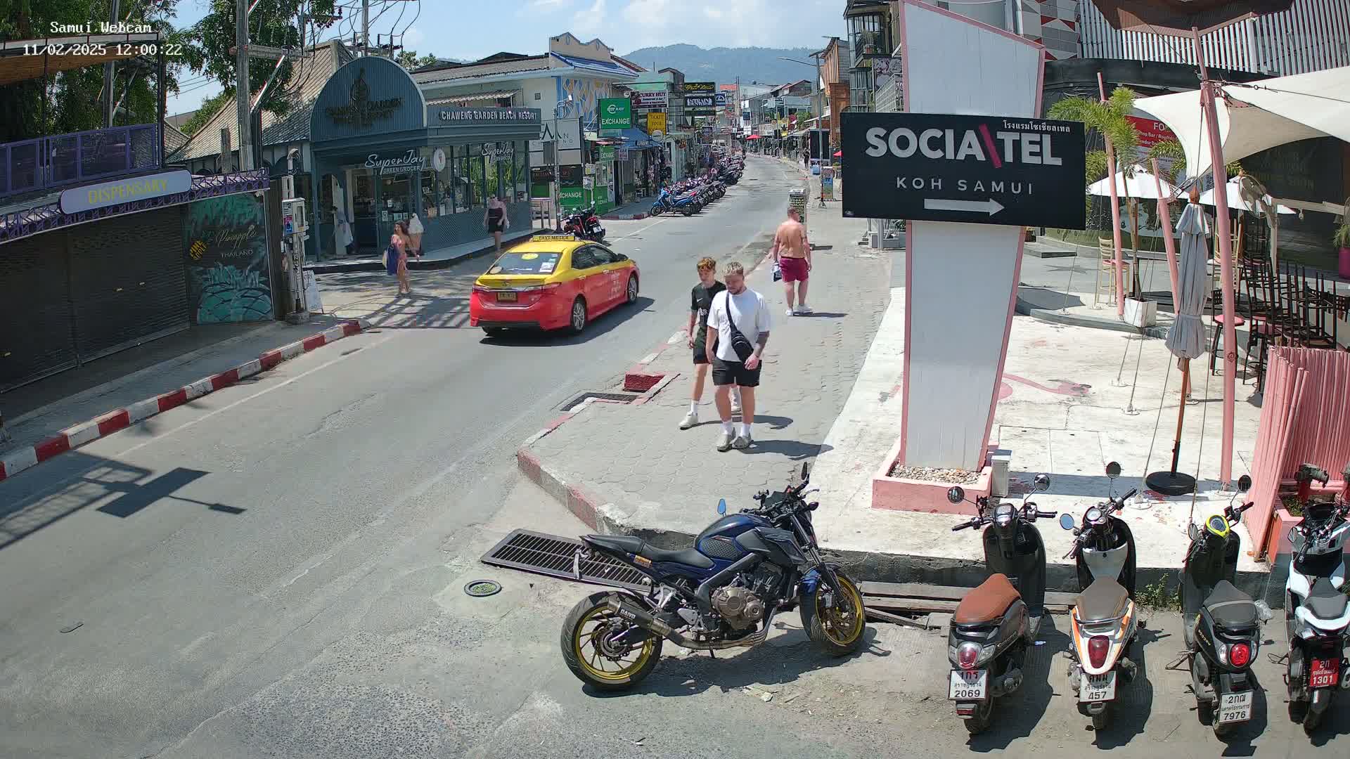 A sunny street scene shows pedestrians and vehicles, including a taxi and several motorbikes parked near a cafe.