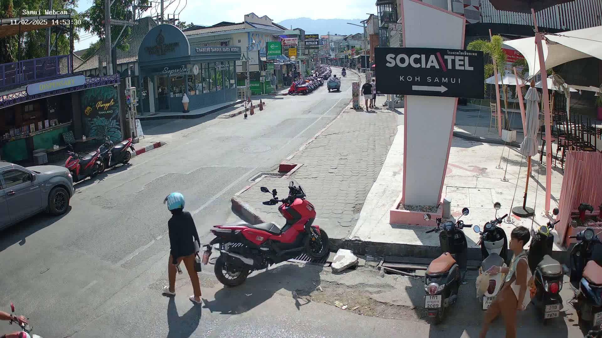 A sunny street scene shows parked motorbikes, pedestrians, and several shops and restaurants along a road lined with buildings.