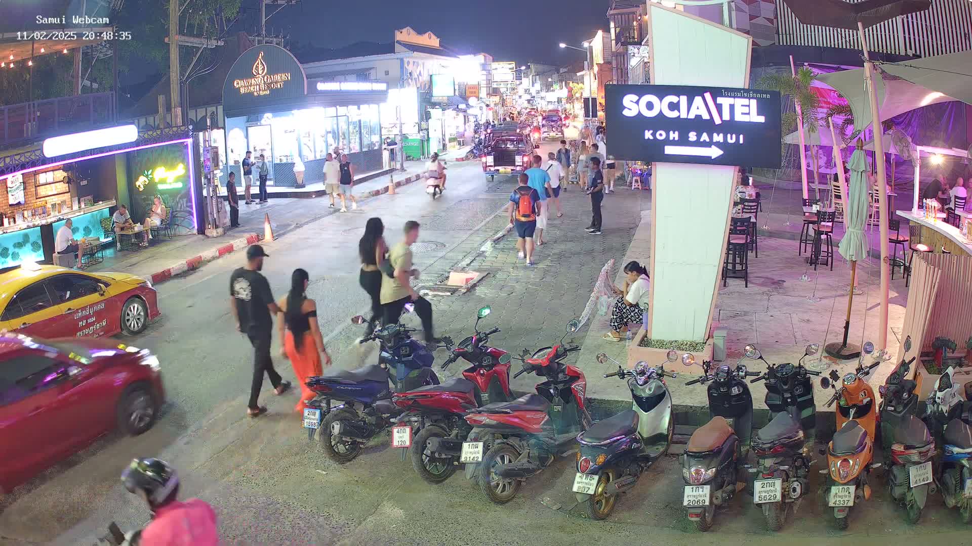 A nighttime street scene shows pedestrians walking among parked motor scooters and cars, under ambient lighting.