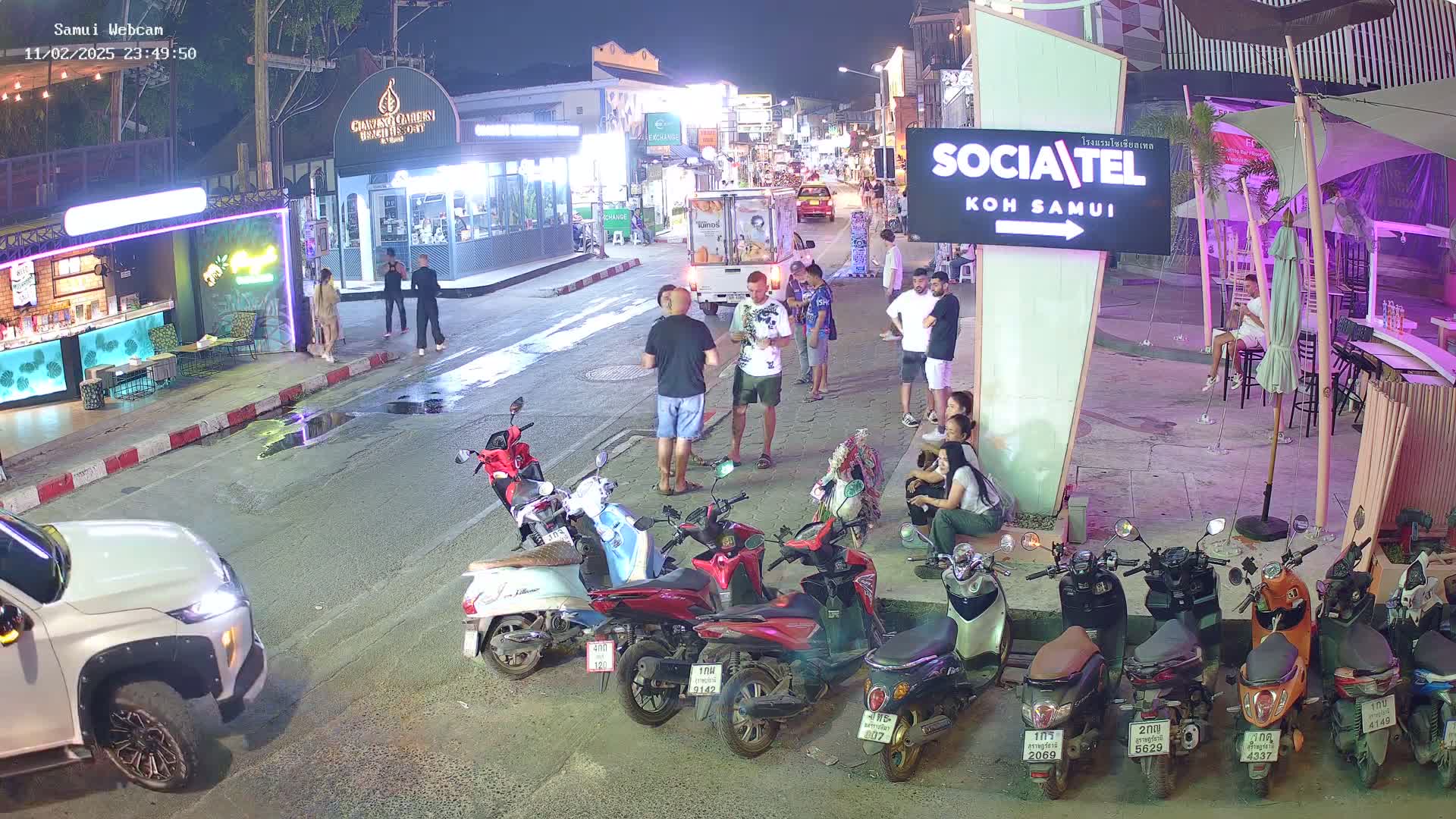 A nighttime street scene shows several people standing and sitting near parked motorbikes and illuminated shops and restaurants.