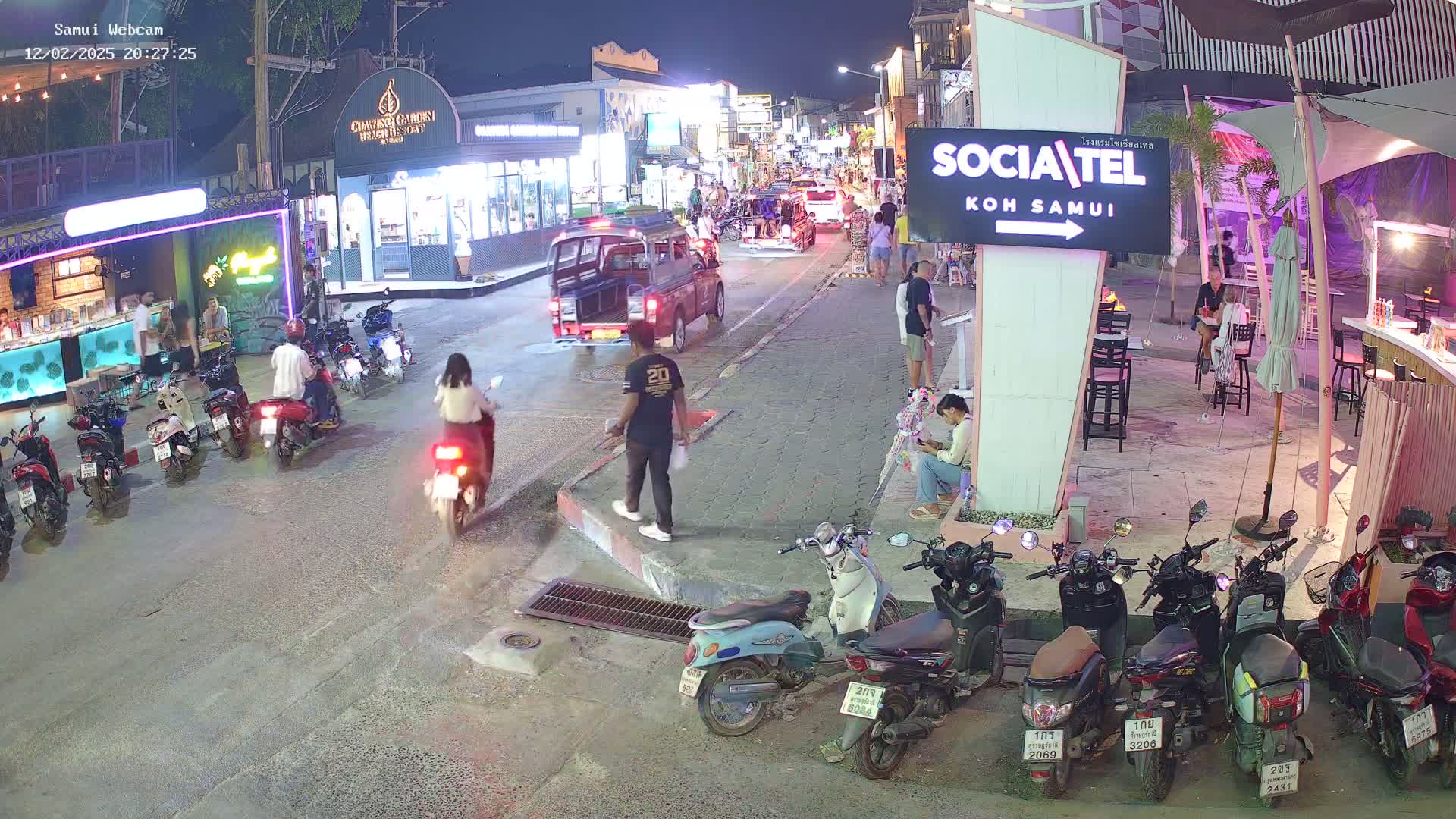 A nighttime street scene shows parked and moving motorbikes, people walking, and several businesses along a street lined with buildings under outdoor lighting.