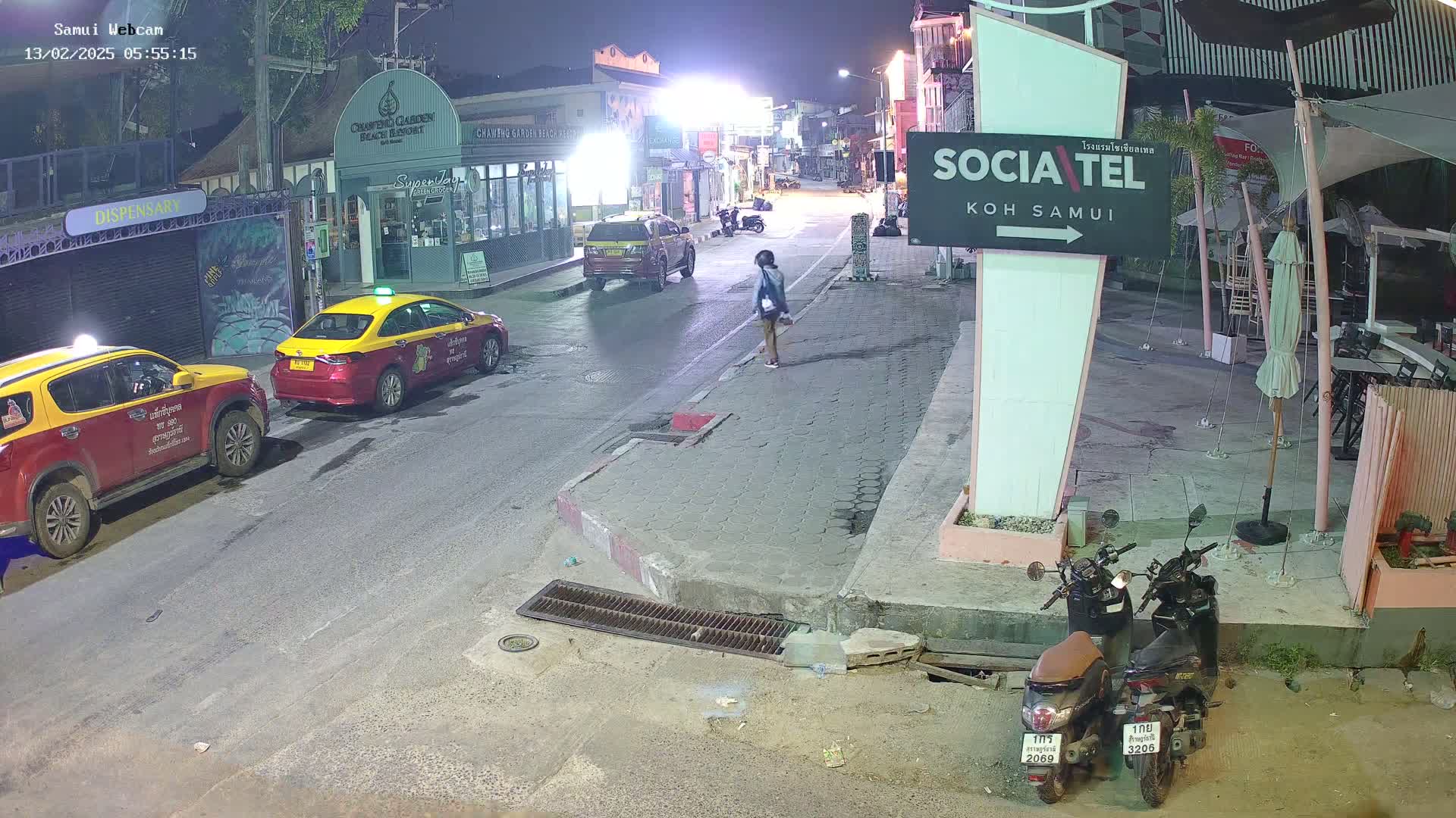 At night, a person walks down a street lined with buildings, past parked taxis and scooters under a clear sky.