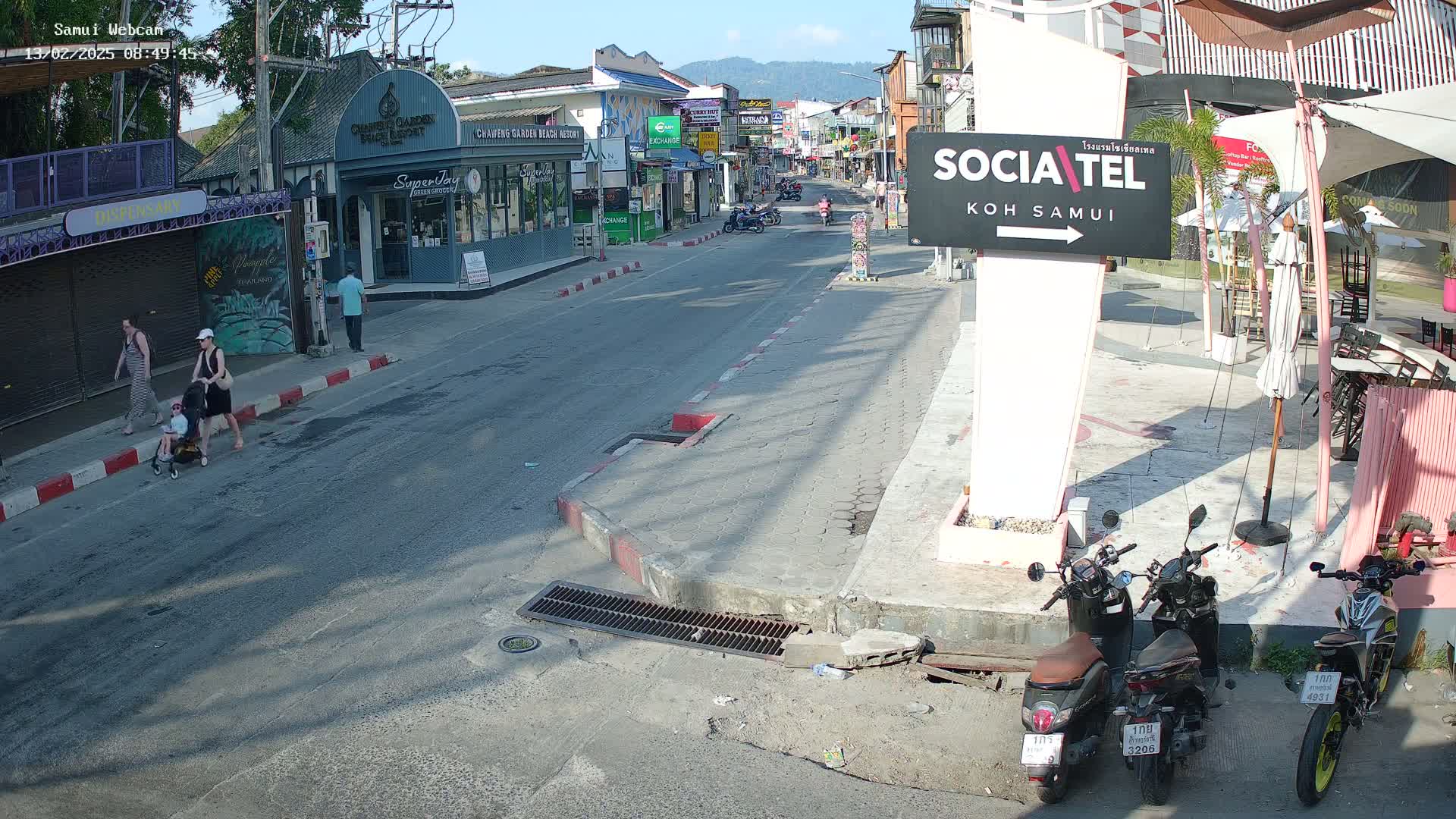 A sunny street scene shows pedestrians, including a woman pushing a stroller, walking along a sidewalk next to a road where several motorbikes are parked, and shops and buildings line the street.