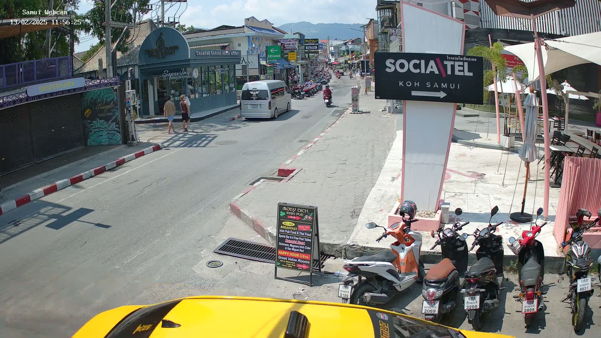 A sunny street scene shows a road with parked motorcycles and a minivan, pedestrians walking on a sidewalk alongside shops and restaurants, and mountains visible in the distance.