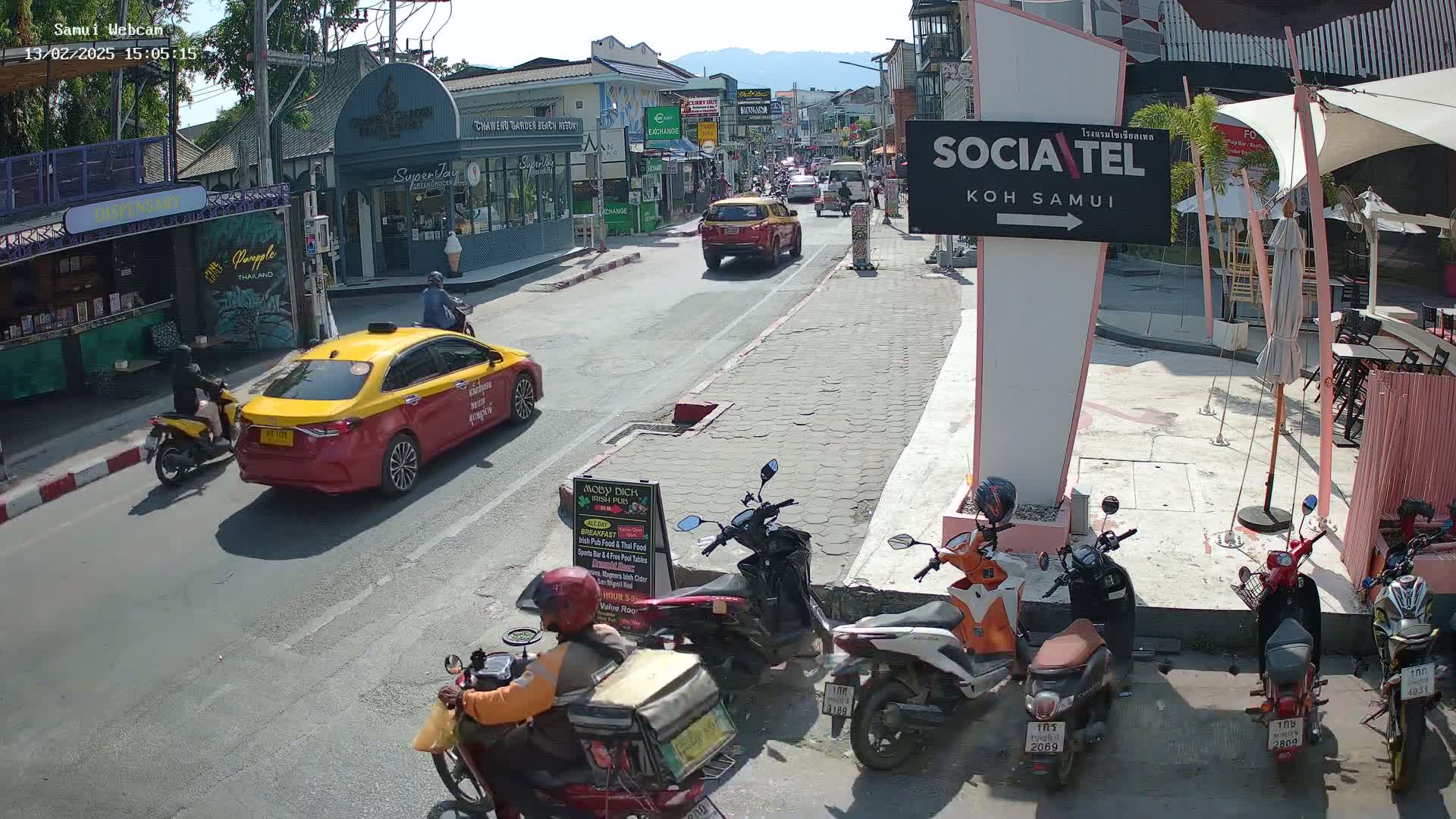 A sunny street scene shows a yellow taxi and several motorbikes parked along the side of a road lined with shops and restaurants, with additional vehicles and pedestrians visible further down the road.