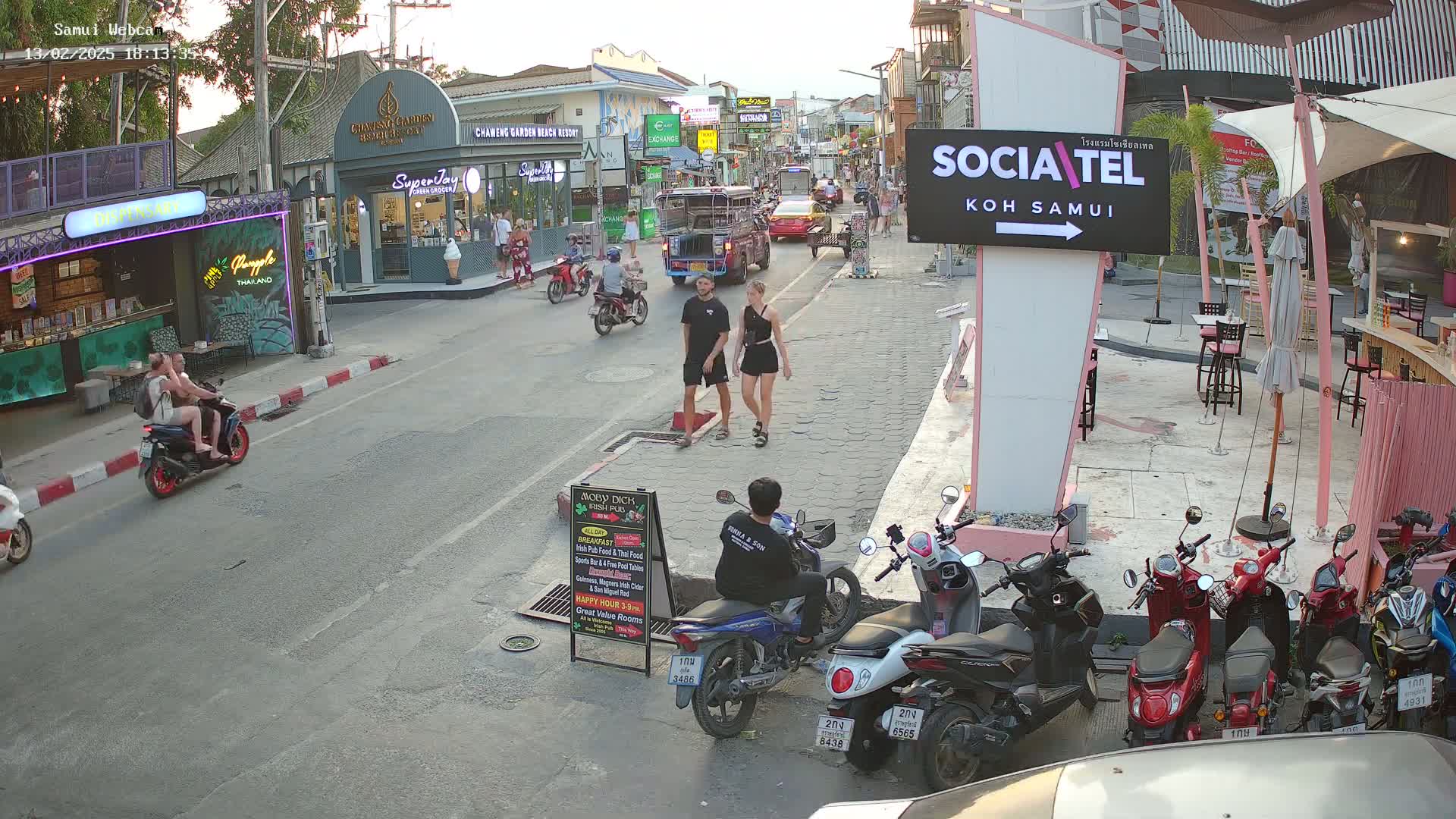 A street scene at dusk shows several people walking and riding motorbikes past shops and restaurants, with parked motorbikes lining the street.
