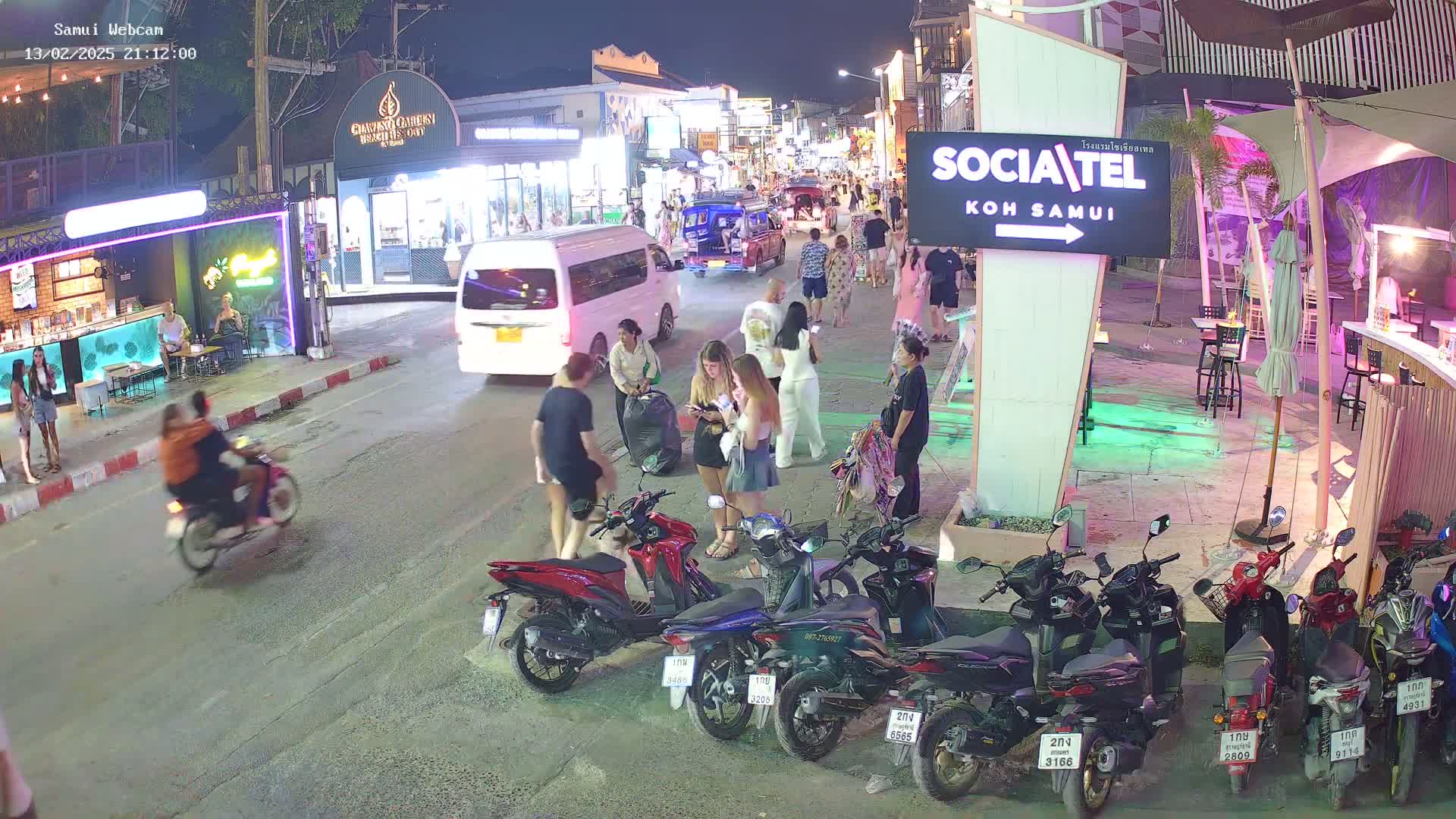 A nighttime street scene in a tropical location shows pedestrians, parked motorbikes, and several vehicles including a van, with a vibrant, brightly lit commercial area in the background.