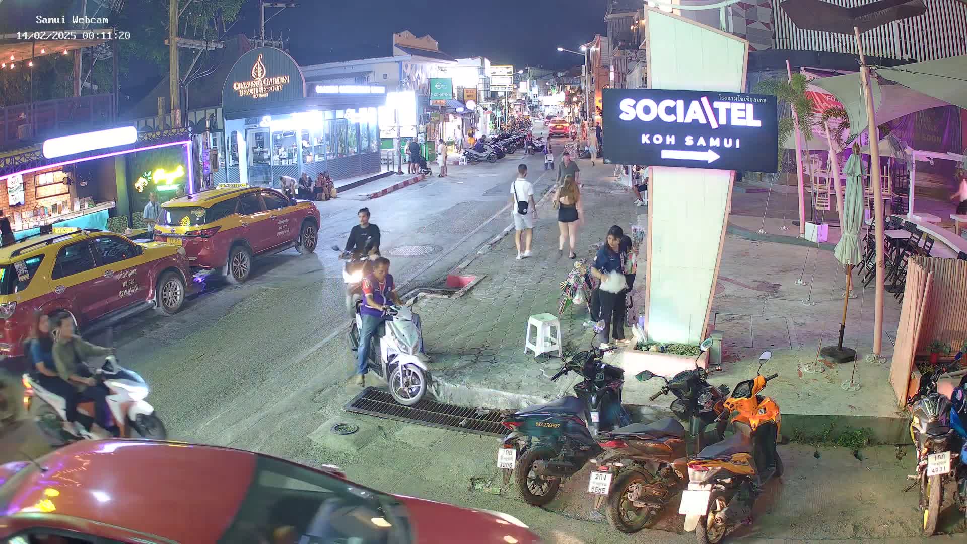 At night, people walk and ride motorbikes on a street lined with shops and businesses, parked motorbikes and taxis are visible.