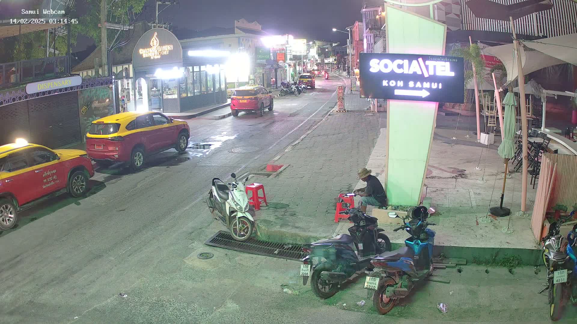 At night, a street scene shows parked cars and motorcycles, a person sitting on the sidewalk, and a sign for a business.