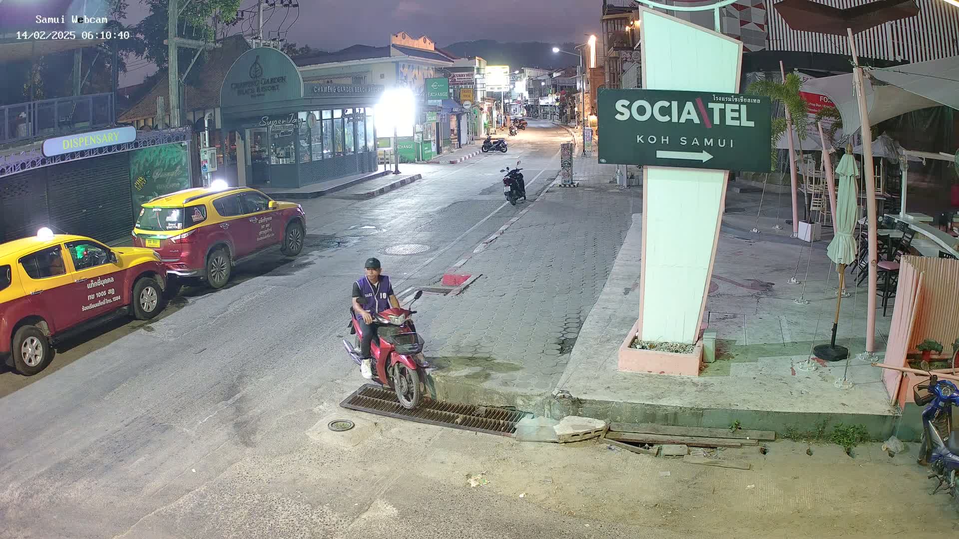 A man on a red motorbike sits near parked taxis on a nighttime street, under a mostly clear sky.