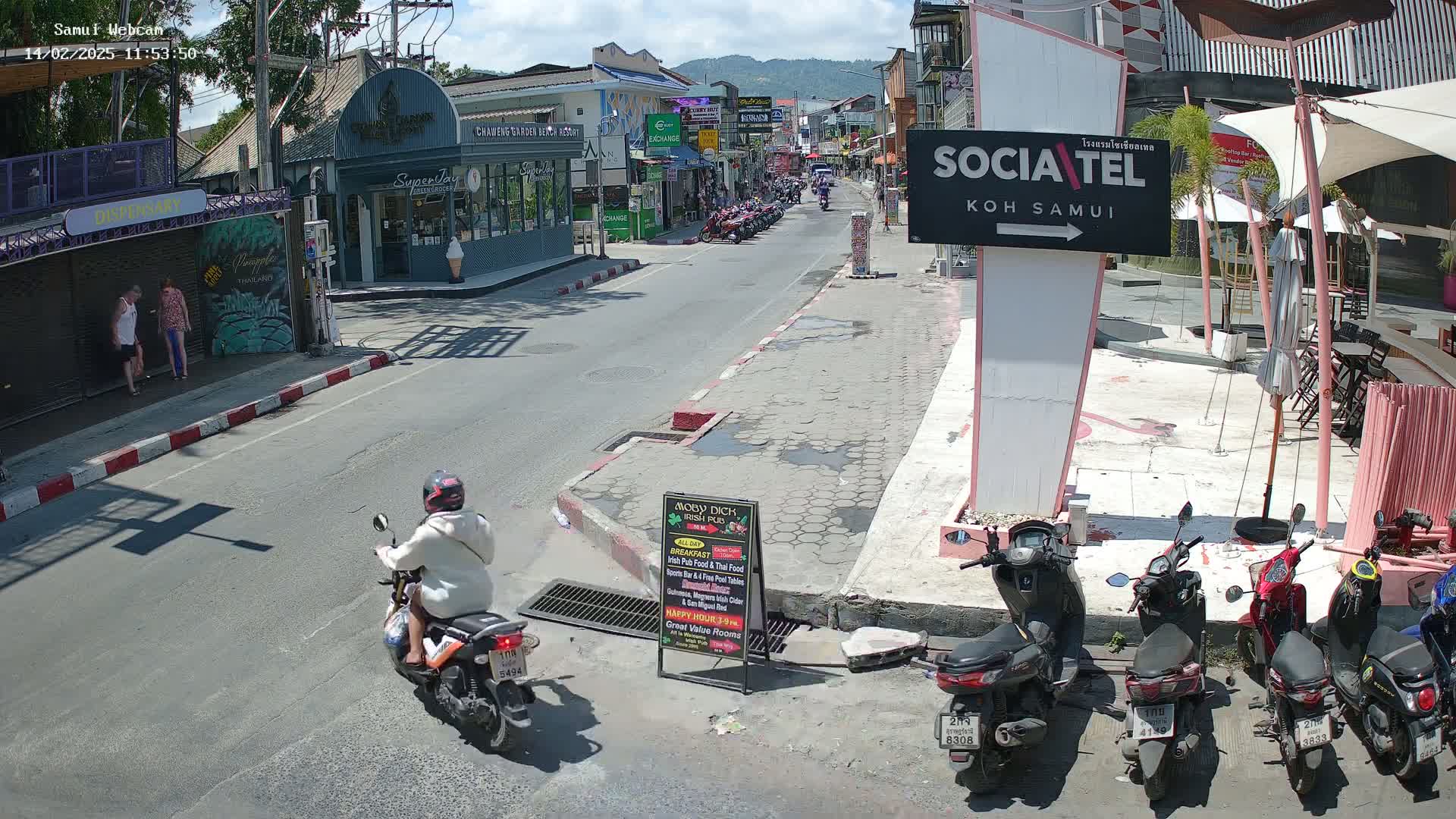 A sunny street scene shows a person on a motor scooter, parked motorcycles, pedestrians, and various shops and restaurants in a tropical location.