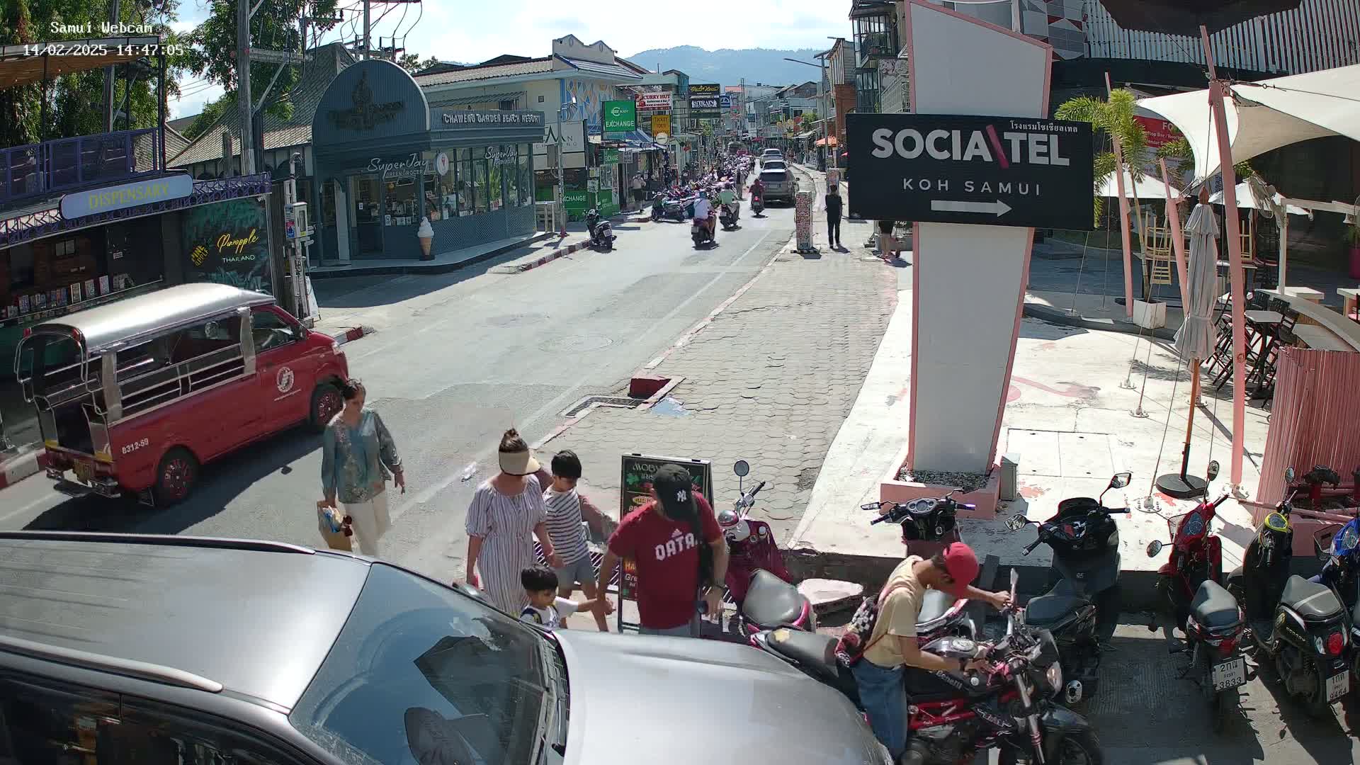 A sunny street scene shows people walking and riding motorbikes and scooters past shops and restaurants, with a red songthaew parked nearby.