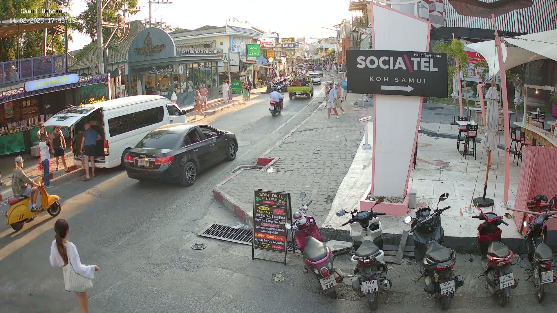 A street scene in bright, late afternoon sunlight shows several cars, scooters, and people, along with shops and restaurants on either side of a road.