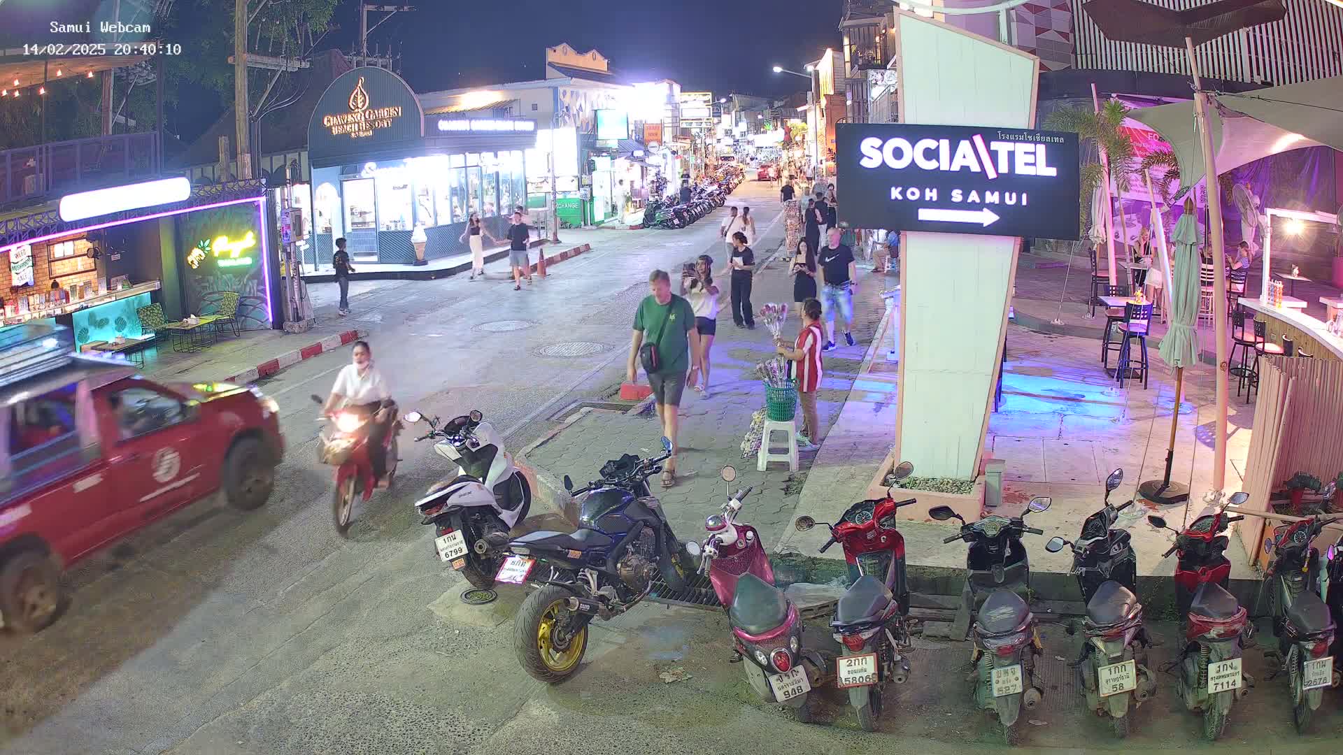 A nighttime street scene shows pedestrians and motorbikes in front of shops and restaurants, under clear skies.
