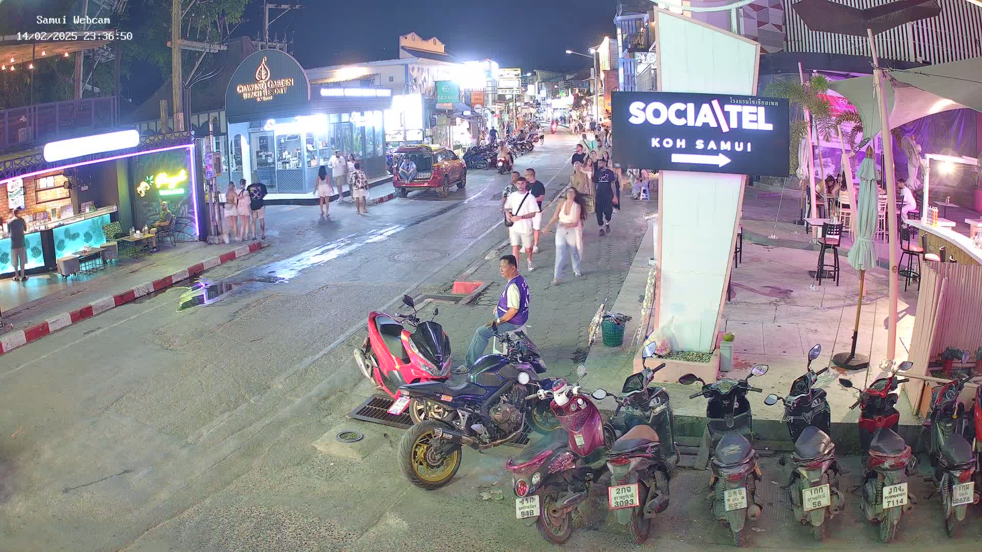 At night, a street scene shows people walking and sitting along a road lined with shops and restaurants, with several parked motor scooters and a man sitting on a motorcycle.