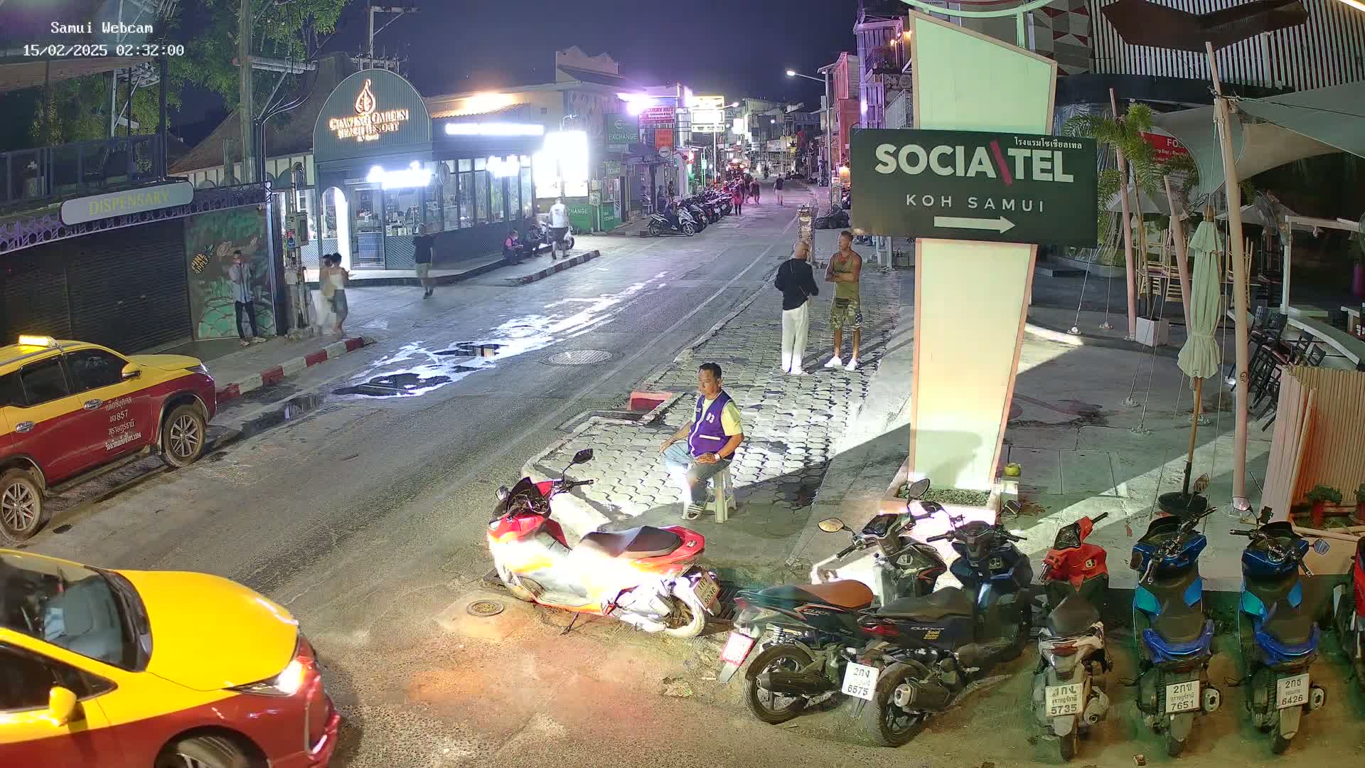 A nighttime street scene shows several people walking and standing near parked motorbikes and cars, with some businesses visible in the background under a clear sky.