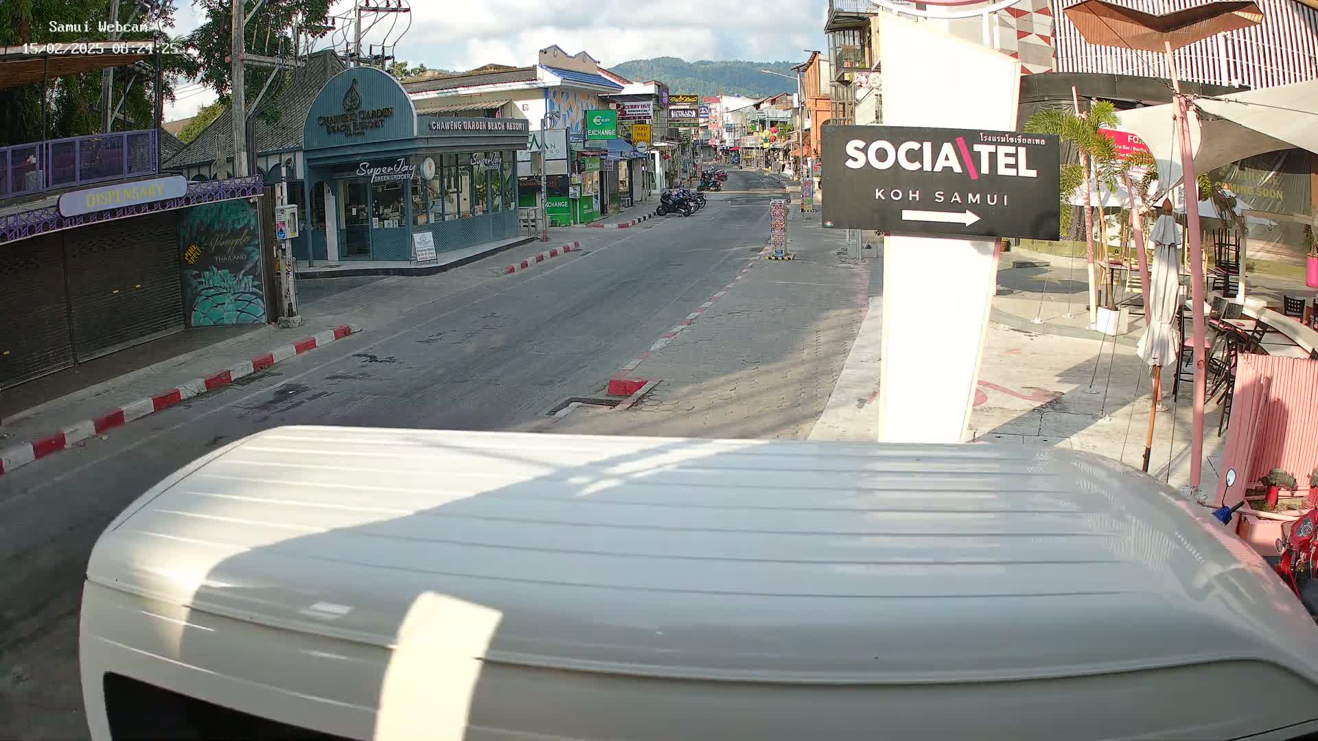 A mostly sunny street scene shows a lightly trafficked road lined with shops and restaurants, viewed from the perspective of a vehicle's roof.