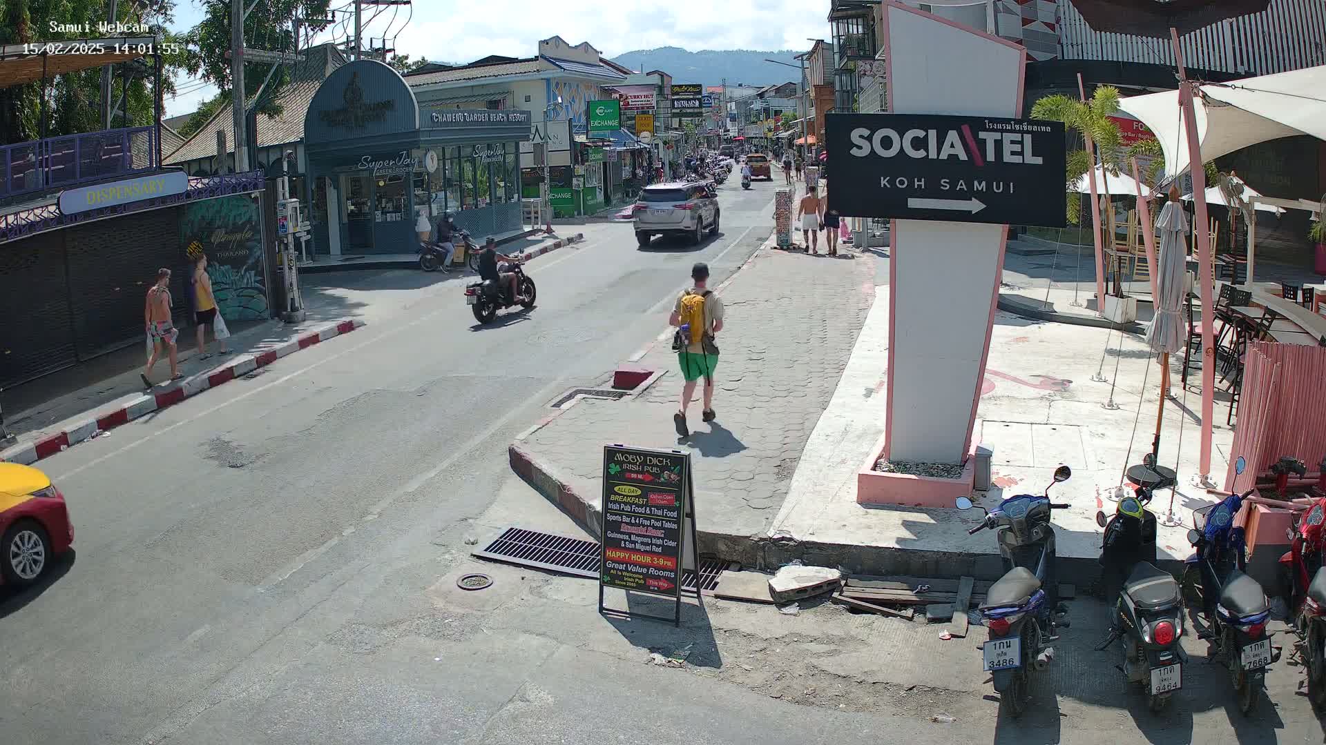A sunny street scene shows pedestrians and motorcyclists on a roadway lined with shops and restaurants, with several parked motorcycles nearby.
